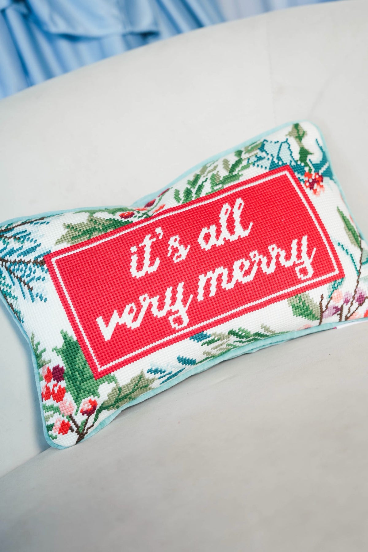 A rectangular decorative pillow with a festive cross-stitch design, featuring a red central panel with the white cursive text 'it's all very merry' and a floral pine border, resting on a light-colored surface.