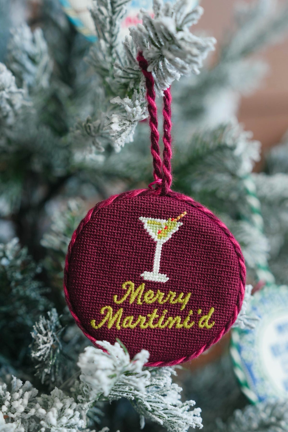 A round embroidered Christmas ornament hanging on a frosted Christmas tree. The ornament is deep burgundy with a yellow and white martini glass design and the text 'Merry Martini'd' in yellow.