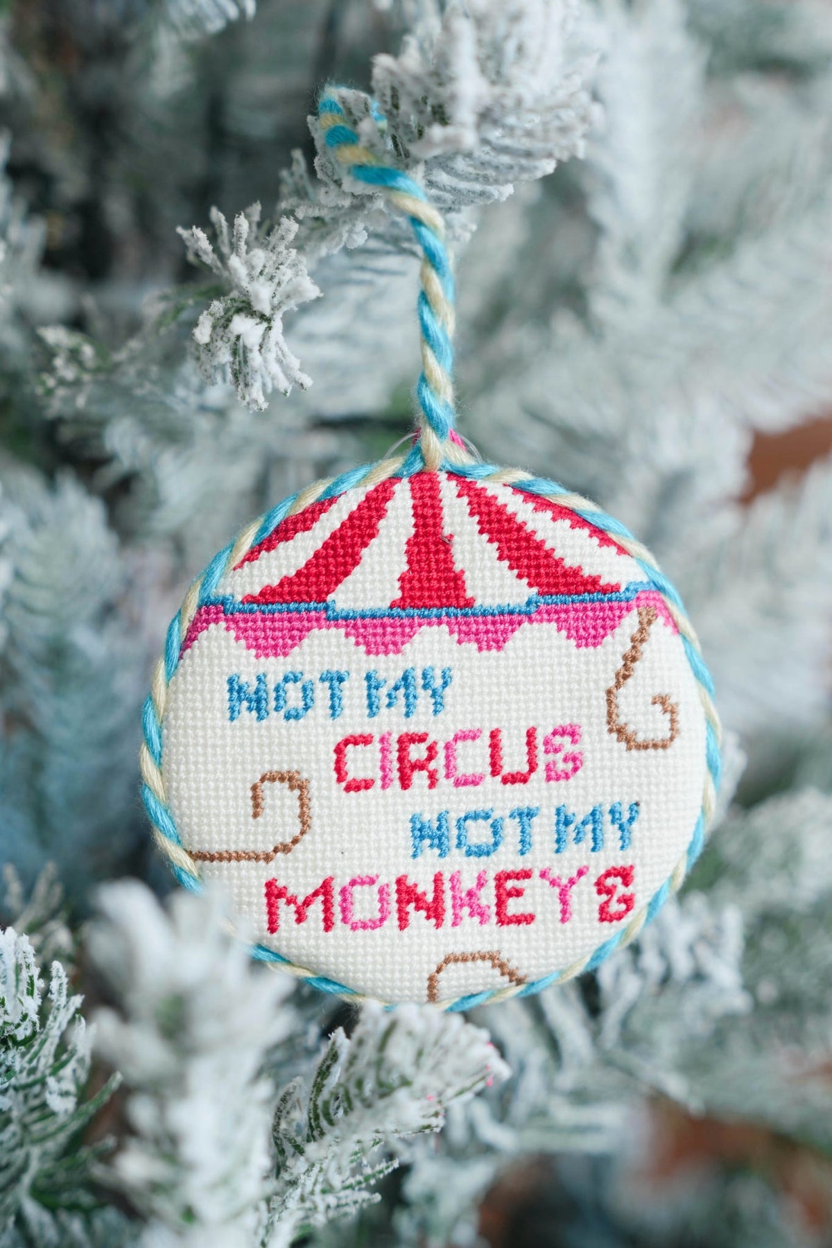 A round embroidered cross-stitch Christmas ornament hanging from a frosted tree branch. The ornament features a red and white striped circus tent at the top and the embroidered text 'NOT MY CIRCUS NOT MY MONKEYS' in blue and red, with small brown monkey silhouettes.