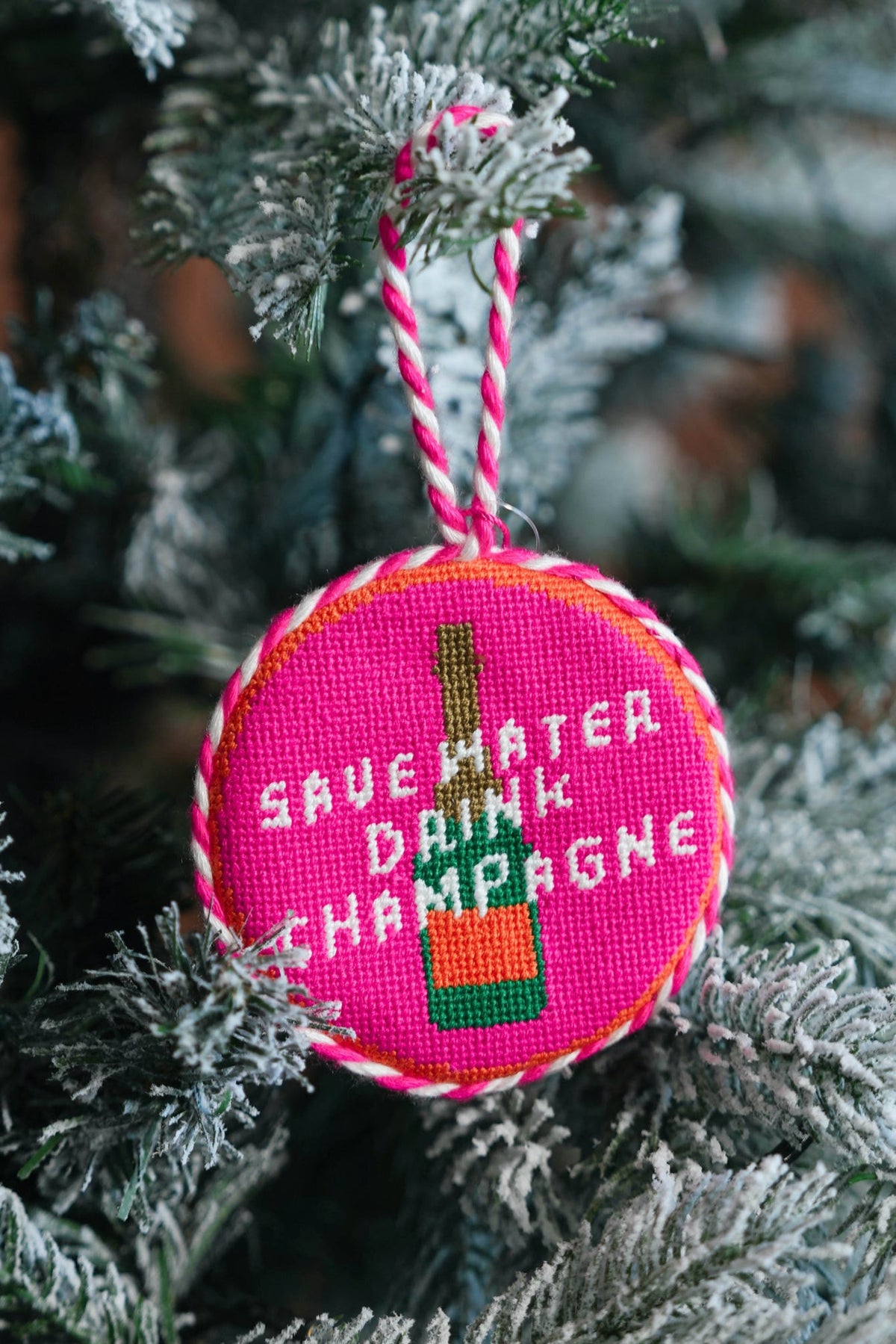 A round embroidered Christmas ornament hanging on a frosted Christmas tree branch. The ornament is bright pink with white text reading 'SAVE WATER DRINK CHAMPAGNE' and a green-and-orange champagne bottle design.