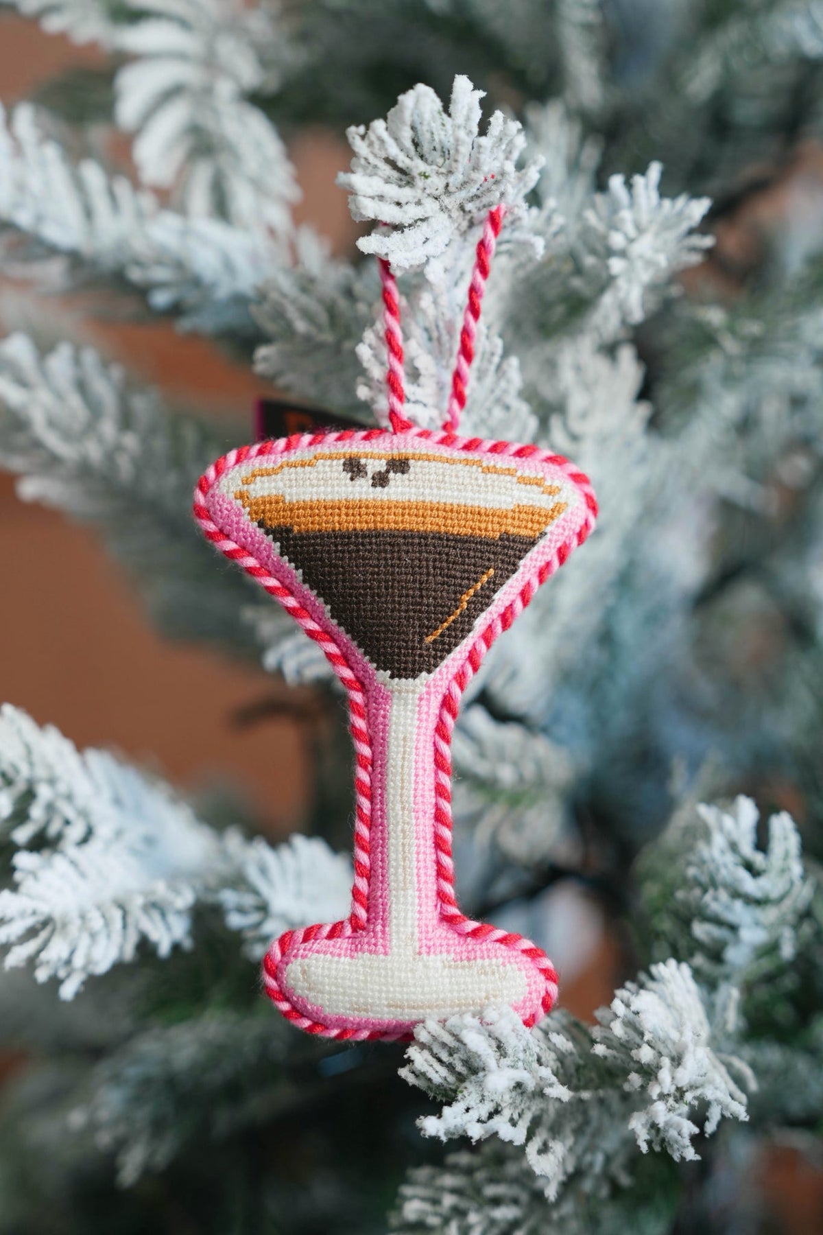 A handmade cross-stitch ornament shaped like a martini glass, hanging on a frosted Christmas tree. The embroidery features a cream-colored body, a pink and white braided border, and a drink design with brown, orange, and black details.