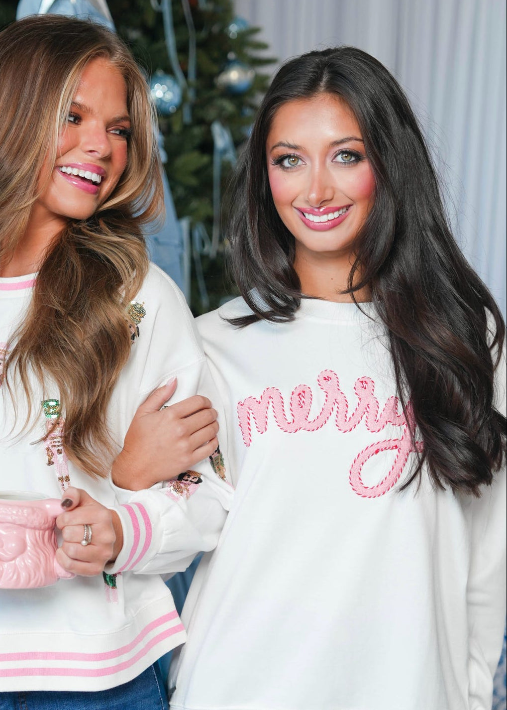 Two women posing together in matching white crewneck sweaters with pink accents. The woman on the left has pink floral embroidery and striped cuffs and hem, holding a pink mug.