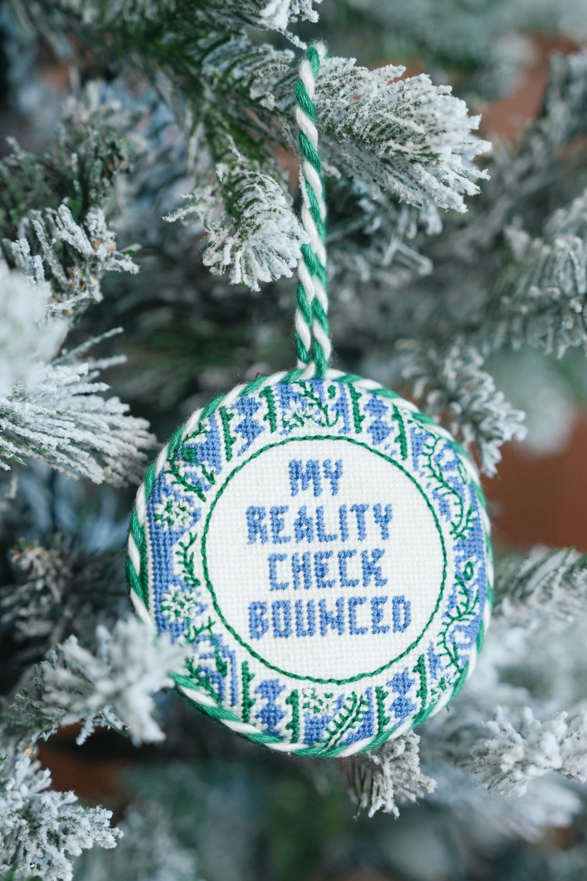 A round cross-stitch Christmas ornament hanging on a frosted tree branch. The ornament features blue stitched text reading 'MY REALITY CHECK BOUNCED' inside a green and blue floral border, suspended by a green and white twisted string.