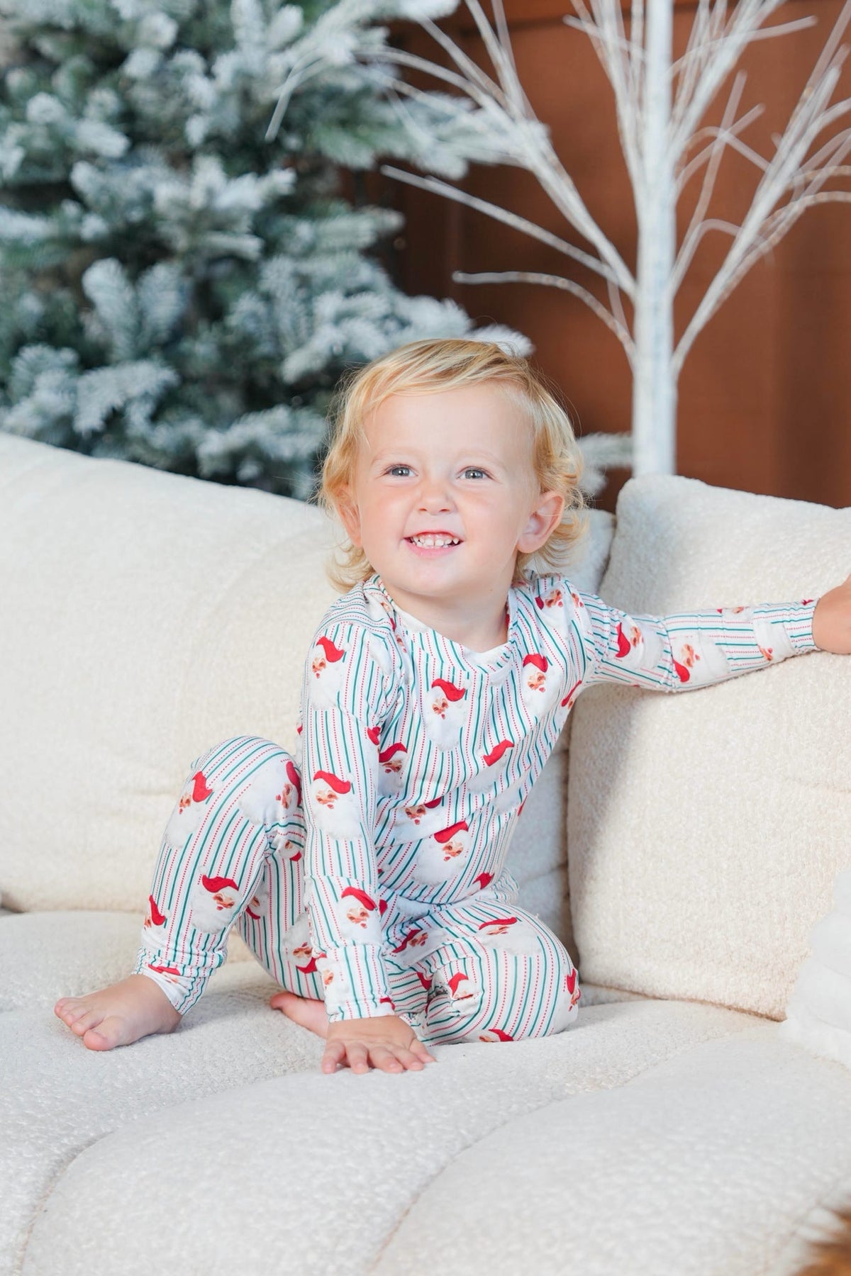 A smiling toddler sits on a cream couch wearing a long-sleeve pajama set with thin blue stripes and red cartoon horse motifs. A blurred Christmas tree and decorative white branches are in the background.