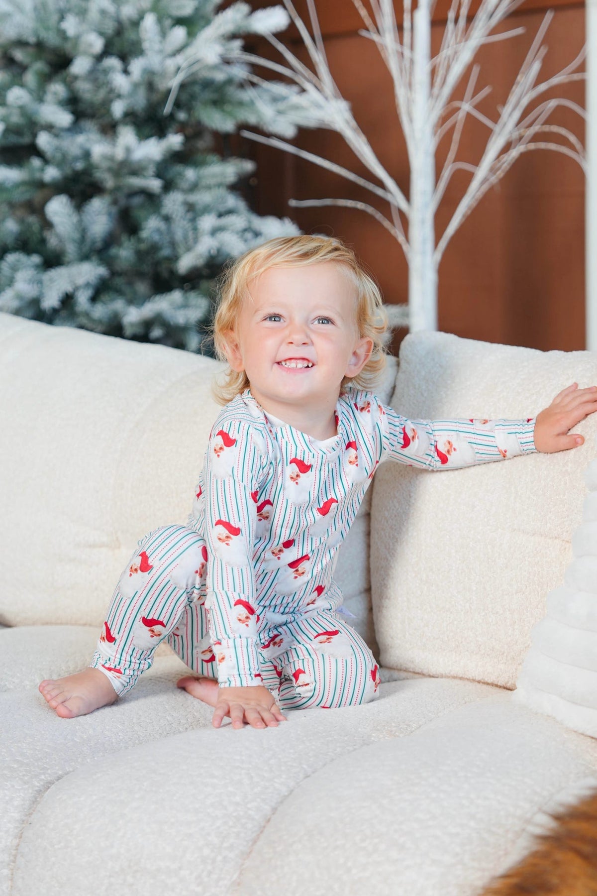 A smiling toddler sits on a cream couch wearing a long-sleeve pajama set with thin blue pinstripes and red bird motifs. The background features a decorated Christmas tree and a white decorative tree branch.
