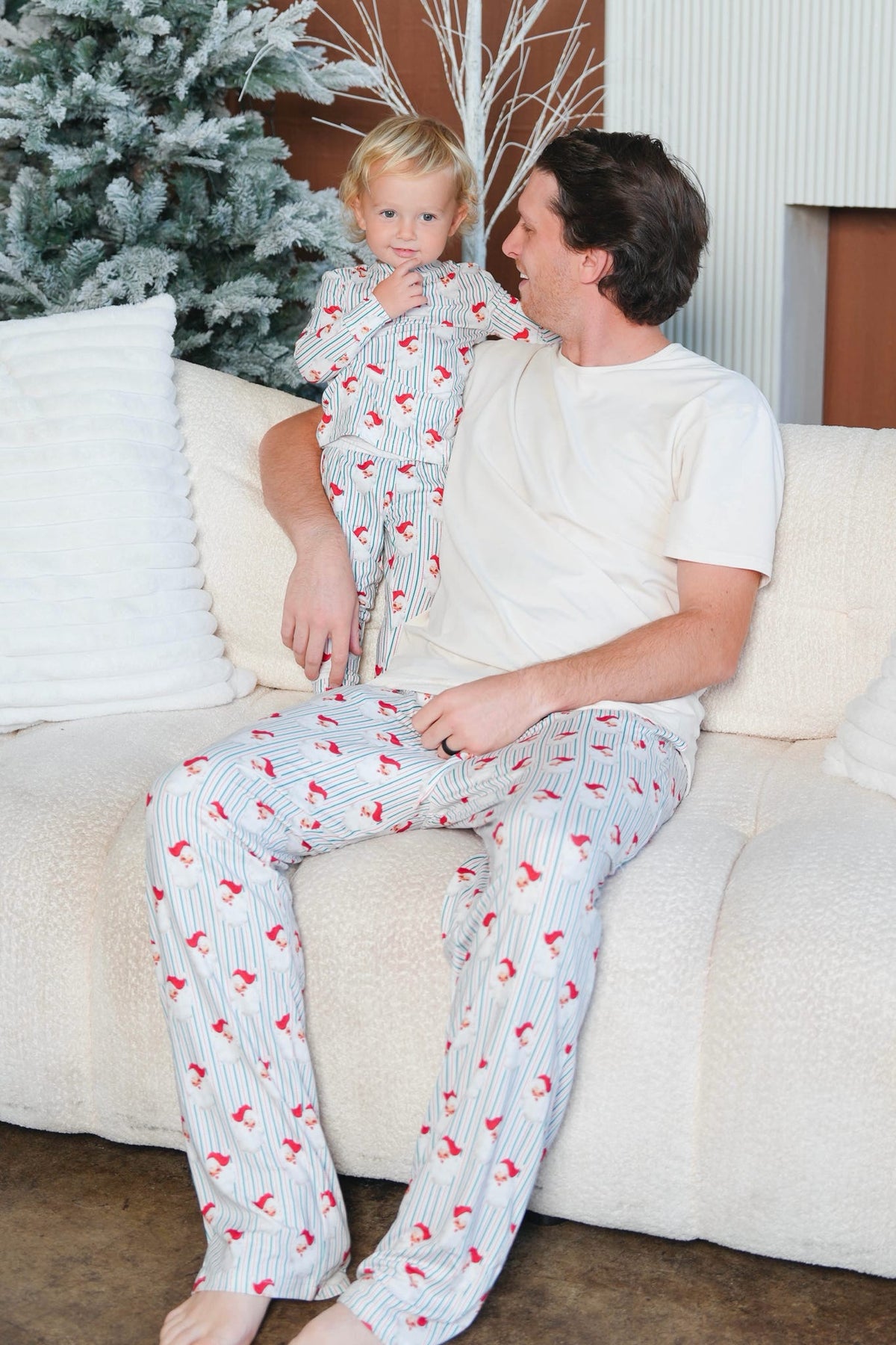 A man and a young child wearing matching holiday-themed pajamas with red birds on a white pinstripe background, sitting together on a cream-colored couch.
