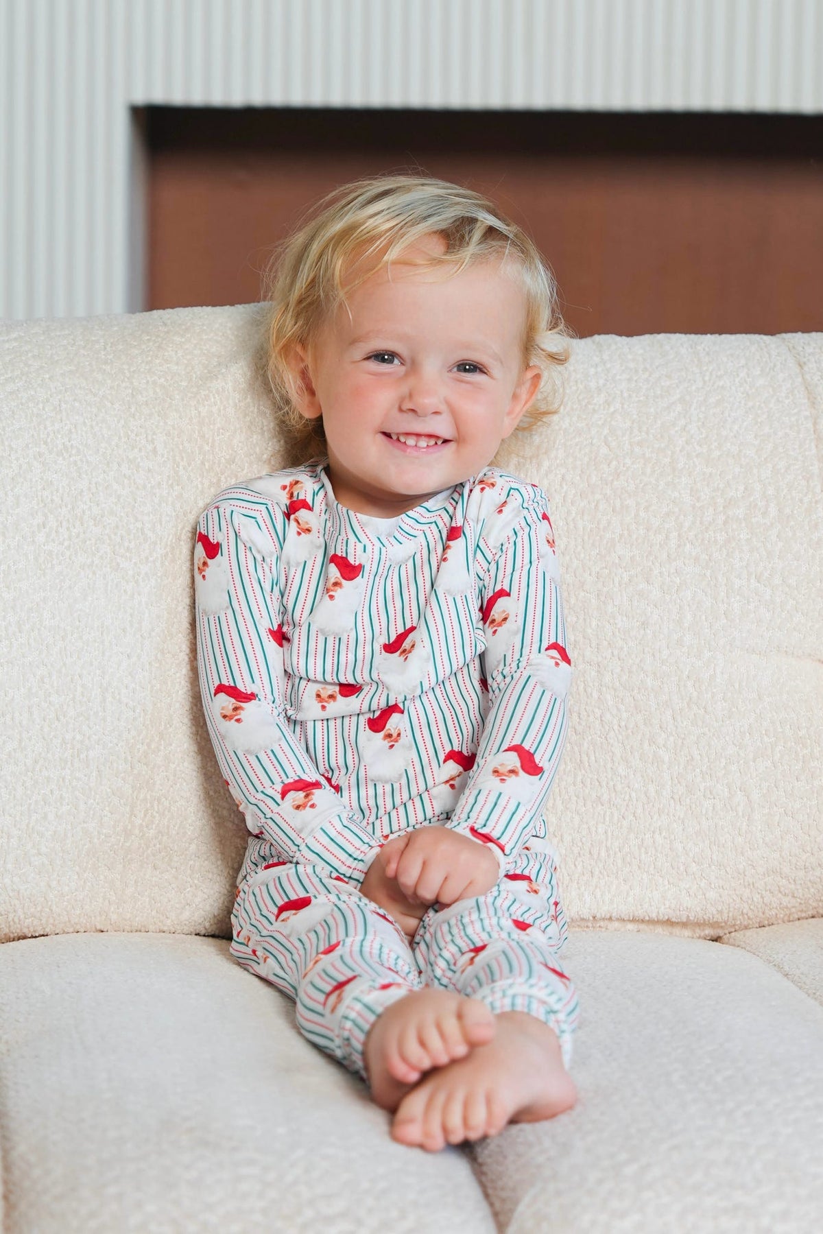 A smiling young child sitting on a cream-colored textured couch, wearing a long-sleeve pajama top and pants set with thin blue vertical stripes and red cartoon foxes.