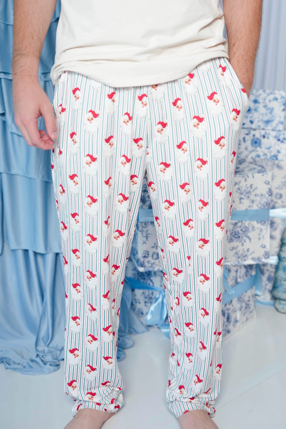 A person wearing white pajama pants with thin blue vertical stripes and a repeating red bird print, paired with a light beige t-shirt. The background shows a blue patterned bed and light blue drapery.