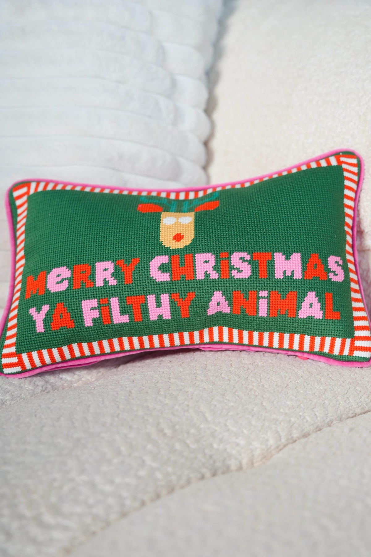 A rectangular Christmas needlepoint pillow with a green background, pink and white striped border, and the text 'MERRY CHRISTMAS YA FILTHY ANIMAL' in pink and red letters, resting on a white textured blanket.