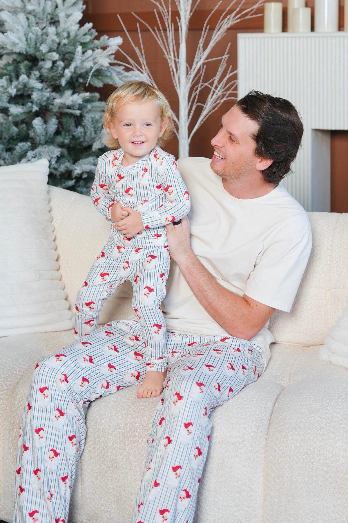 A man and a young child wearing matching pajamas with thin red pinstripes and small red bird motifs, sitting together on a cream-colored couch in a cozy living room setting.