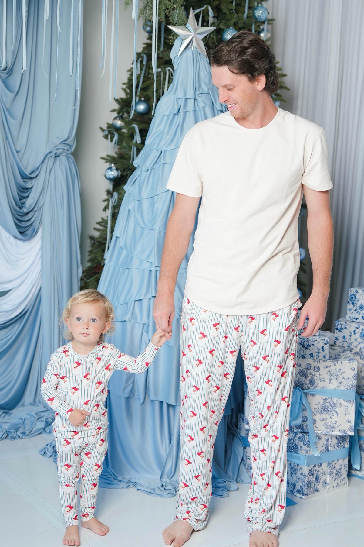 A man and a young child wearing matching white pajama sets with red and blue flamingo prints, standing in a decorated Christmas-themed room with blue drapery and a tree.
