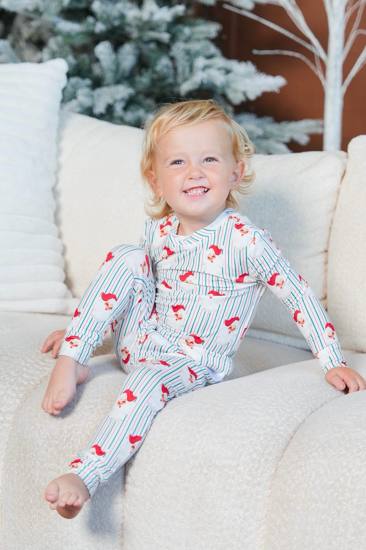 A smiling toddler sits on a cream-colored couch wearing a long-sleeve pajama set with thin blue vertical stripes and red Santa Claus motifs. The background features a cozy holiday scene with a decorated Christmas tree and warm lighting.
