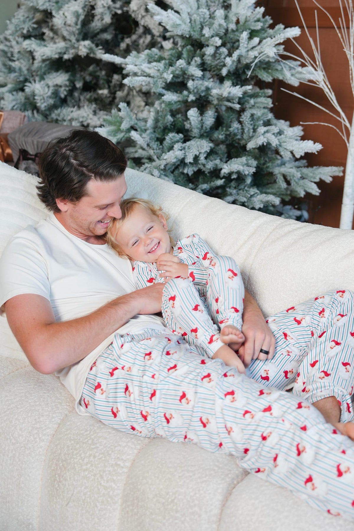 A father and child wearing matching holiday pajamas with a red and white pattern, lying together on a cream-colored couch in front of a decorated Christmas tree.