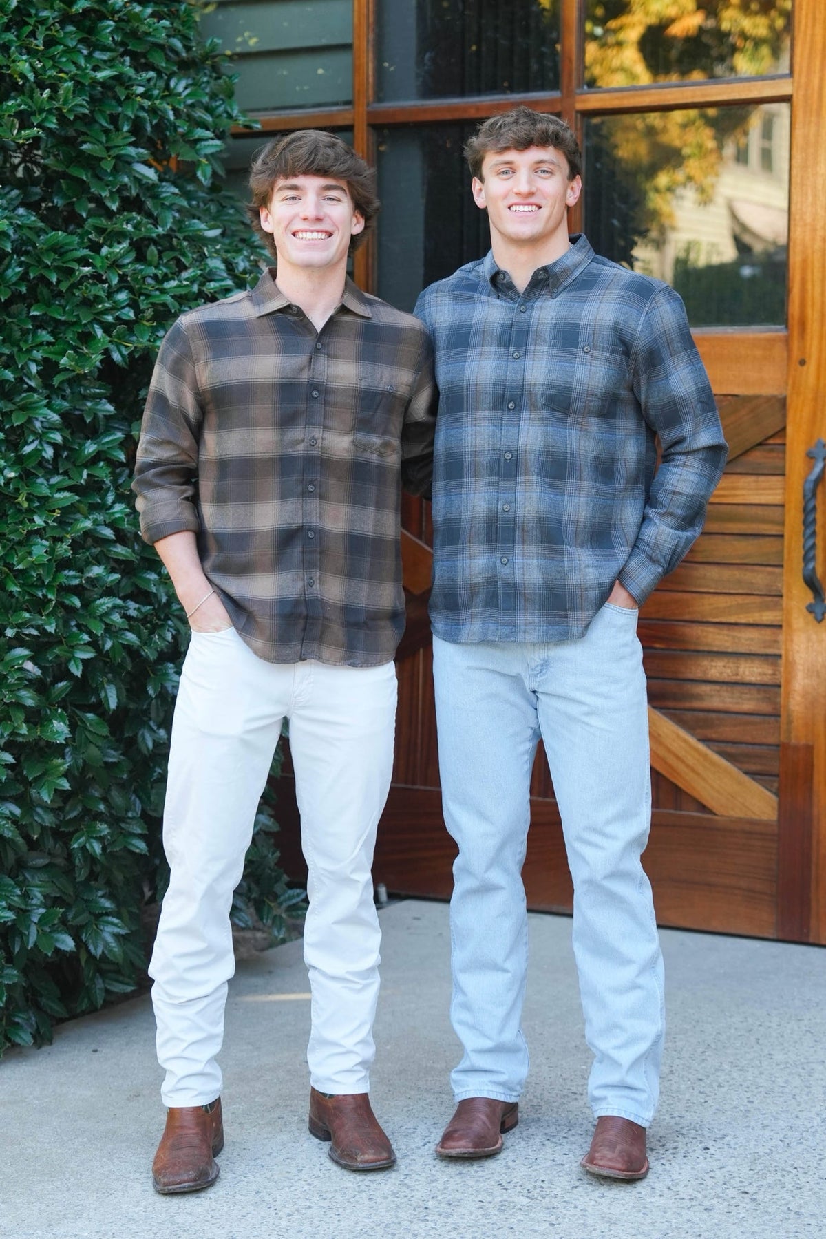 Two young men standing outdoors in front of a wooden door and green ivy, wearing long-sleeve plaid button-up shirts. The man on the left wears a brown and gray plaid shirt with off-white pants, and the man on the right wears a blue and gray plaid shirt with light blue pants.