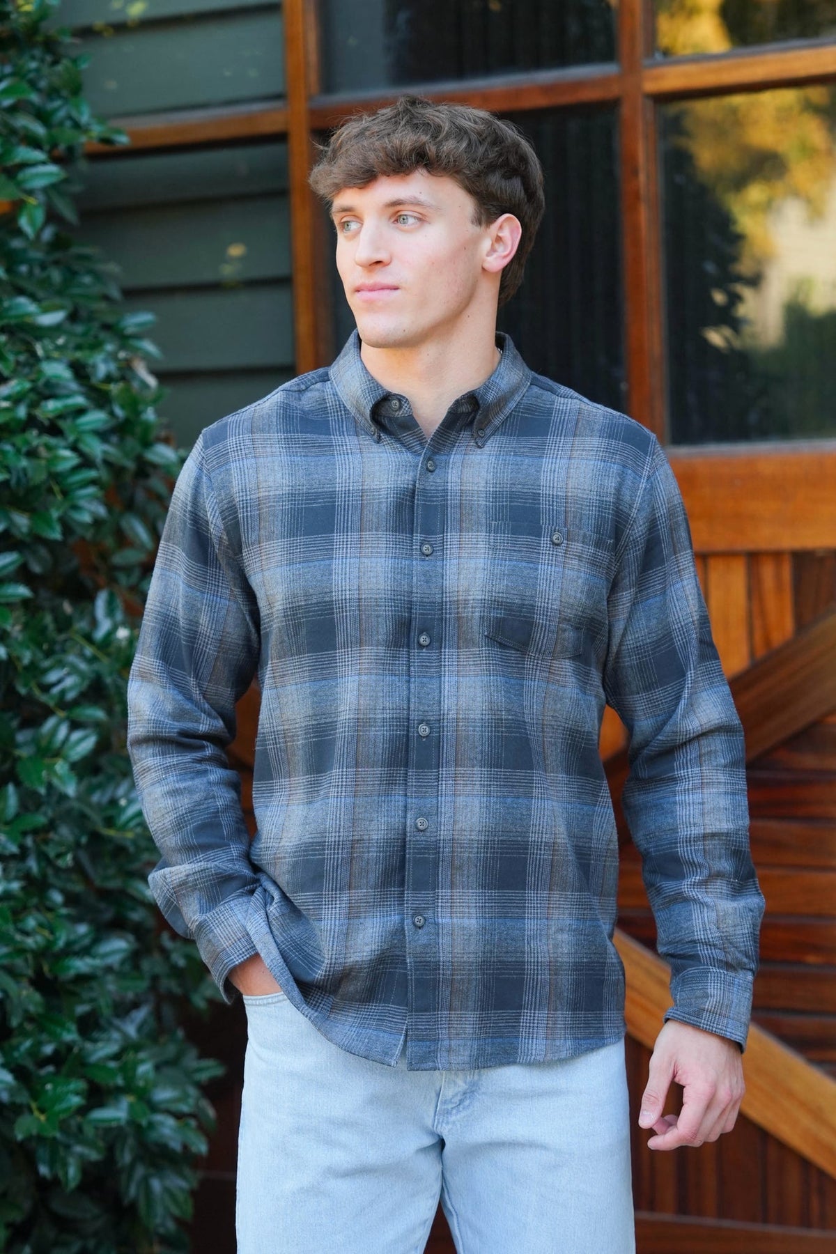 Man wearing a blue and navy blue plaid button down shirt and jeans standing in front of a brown barn door and green bushes. 