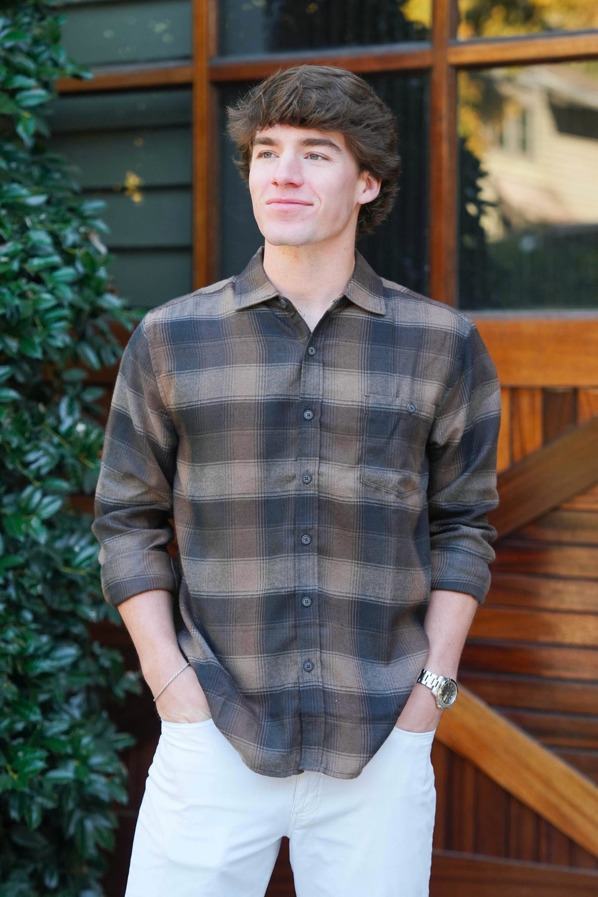 Man wearing a brown and beige plaid button down shirt and jeans standing in front of a brown barn door and green bushes. 