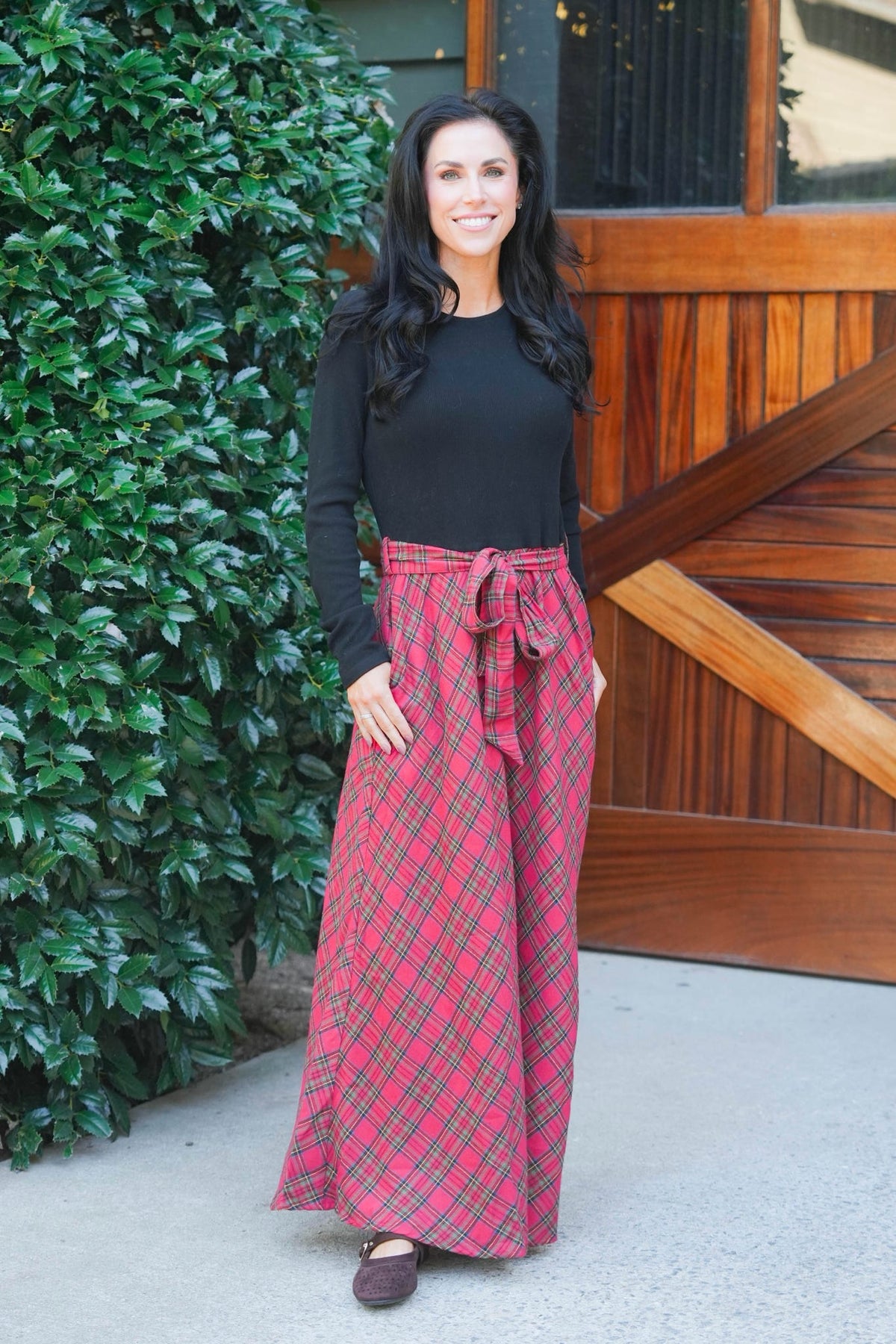 A woman stands outdoors in front of a wooden barn door and green hedge, wearing a black ribbed long-sleeve top and a long plaid skirt with pockets and a matching tie belt.