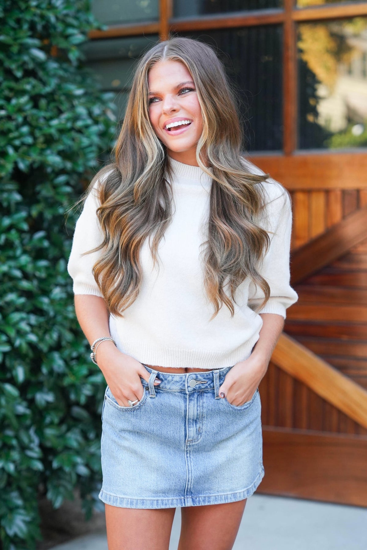 A woman with long wavy brown hair is smiling and looking to the side, wearing a cream-colored mock neck knit sweater and a light-wash denim mini skirt. She is standing outdoors in front of a wooden door and green foliage.