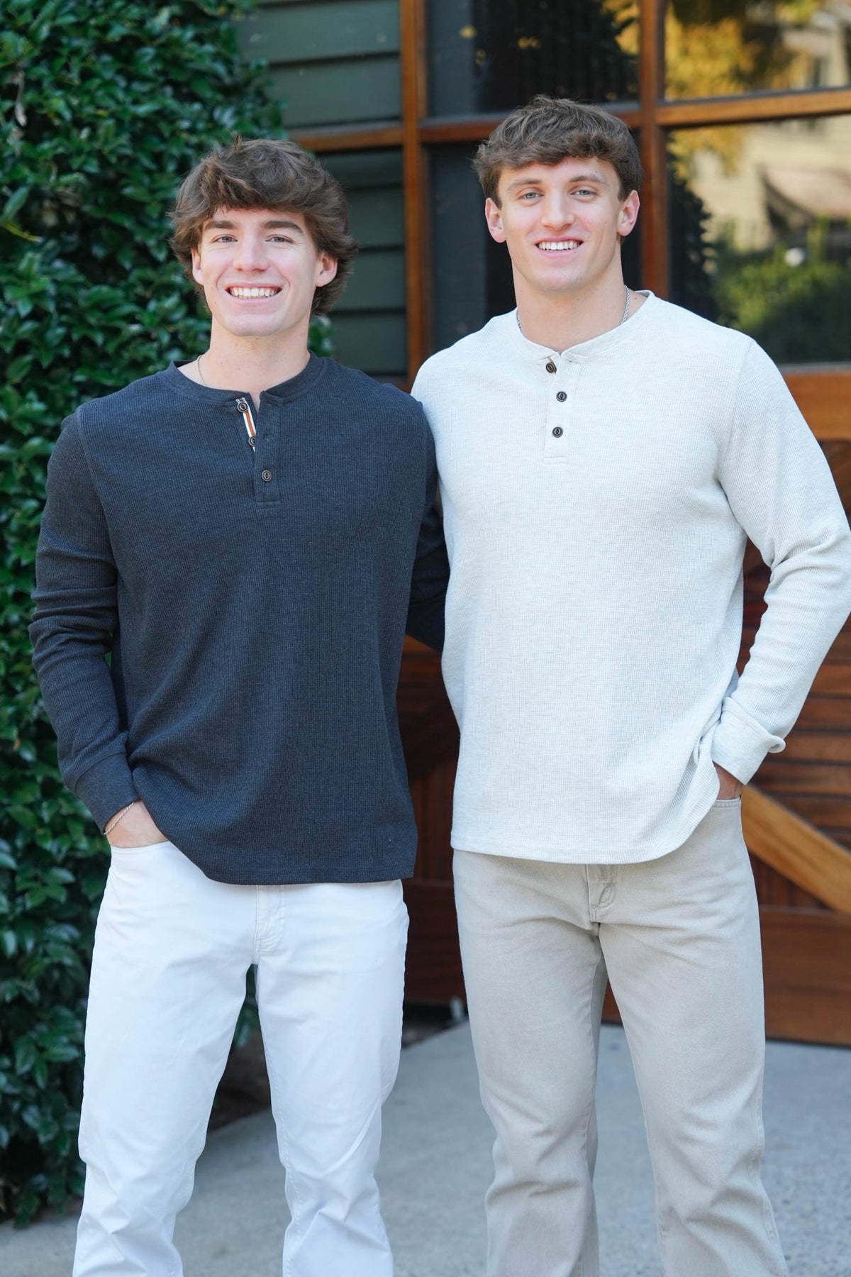 Two young men standing side-by-side outdoors, wearing long-sleeve henley shirts. The man on the left wears a dark charcoal gray henley with white pants, and the man on the right wears an off-white/cream henley with beige pants.