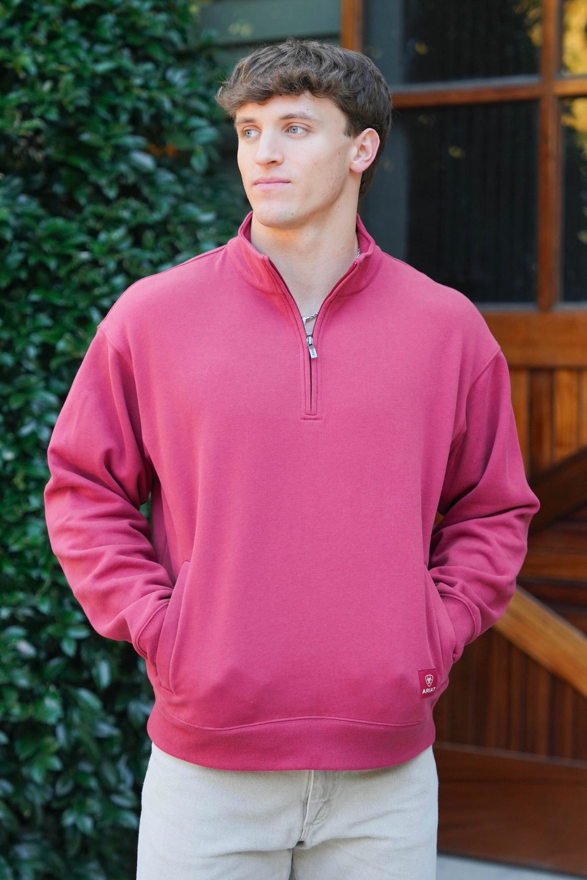A man wearing a dusty rose pink quarter-zip sweatshirt and light beige pants, standing outdoors in front of a wooden door and green foliage.