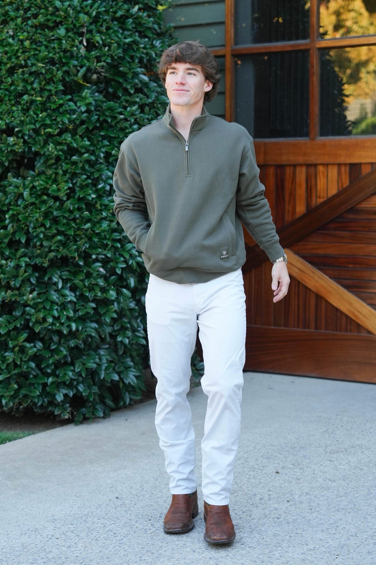 A man wearing an olive green quarter-zip pullover sweater, white pants, and brown boots standing outdoors in front of a wooden barn door and green hedge.