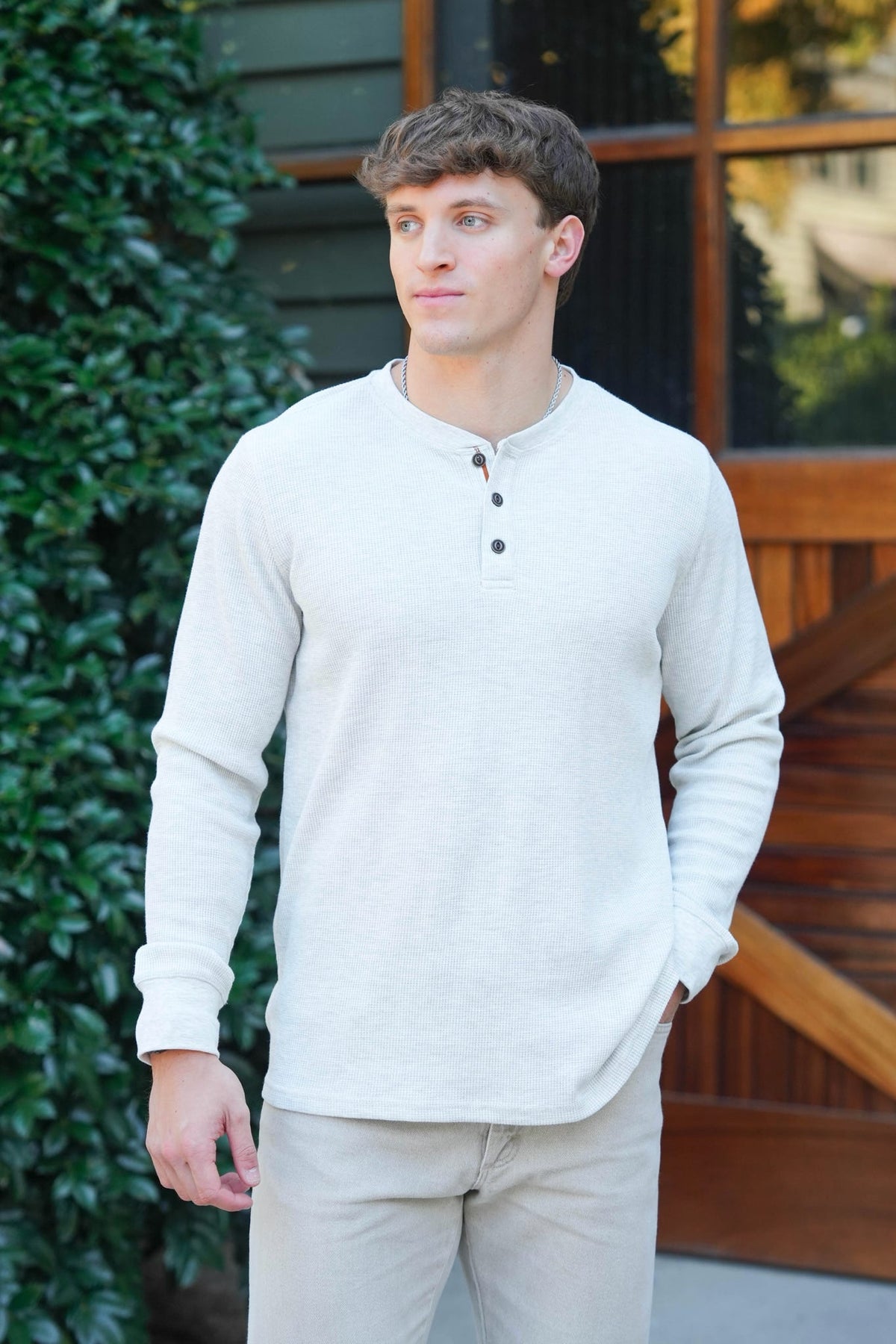 A man wearing a light beige long-sleeve henley shirt and matching beige pants, standing outdoors in front of a wooden building with greenery.