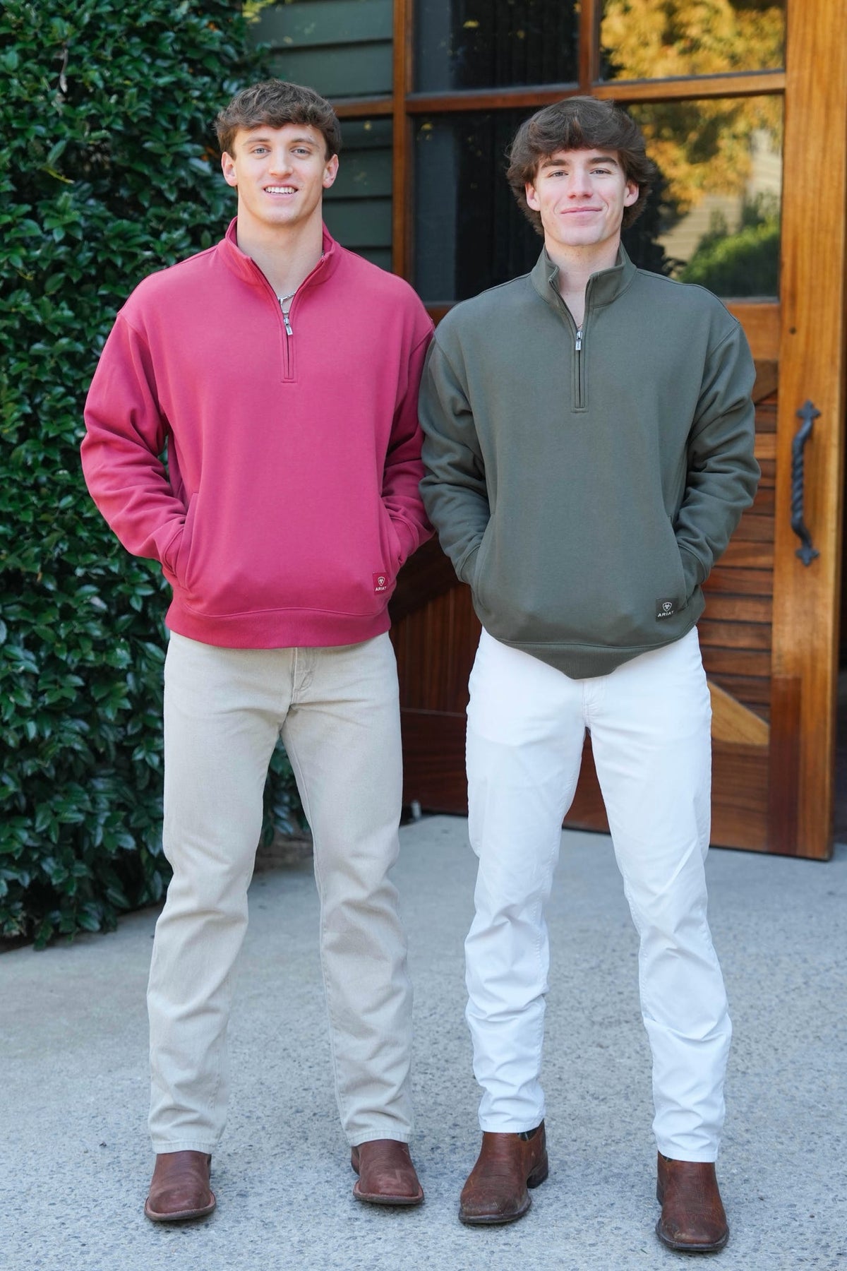 Two young men standing outdoors in front of a wooden door and greenery, wearing quarter-zip pullovers in pink and olive green with light pants and brown boots.