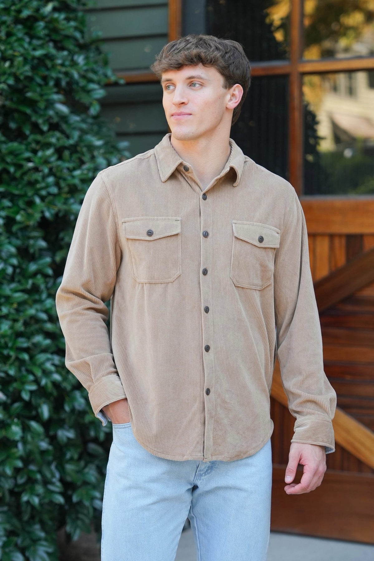 A man wearing a tan corduroy button-down shirt with two chest pockets and light blue jeans, standing outdoors in front of a wooden building.