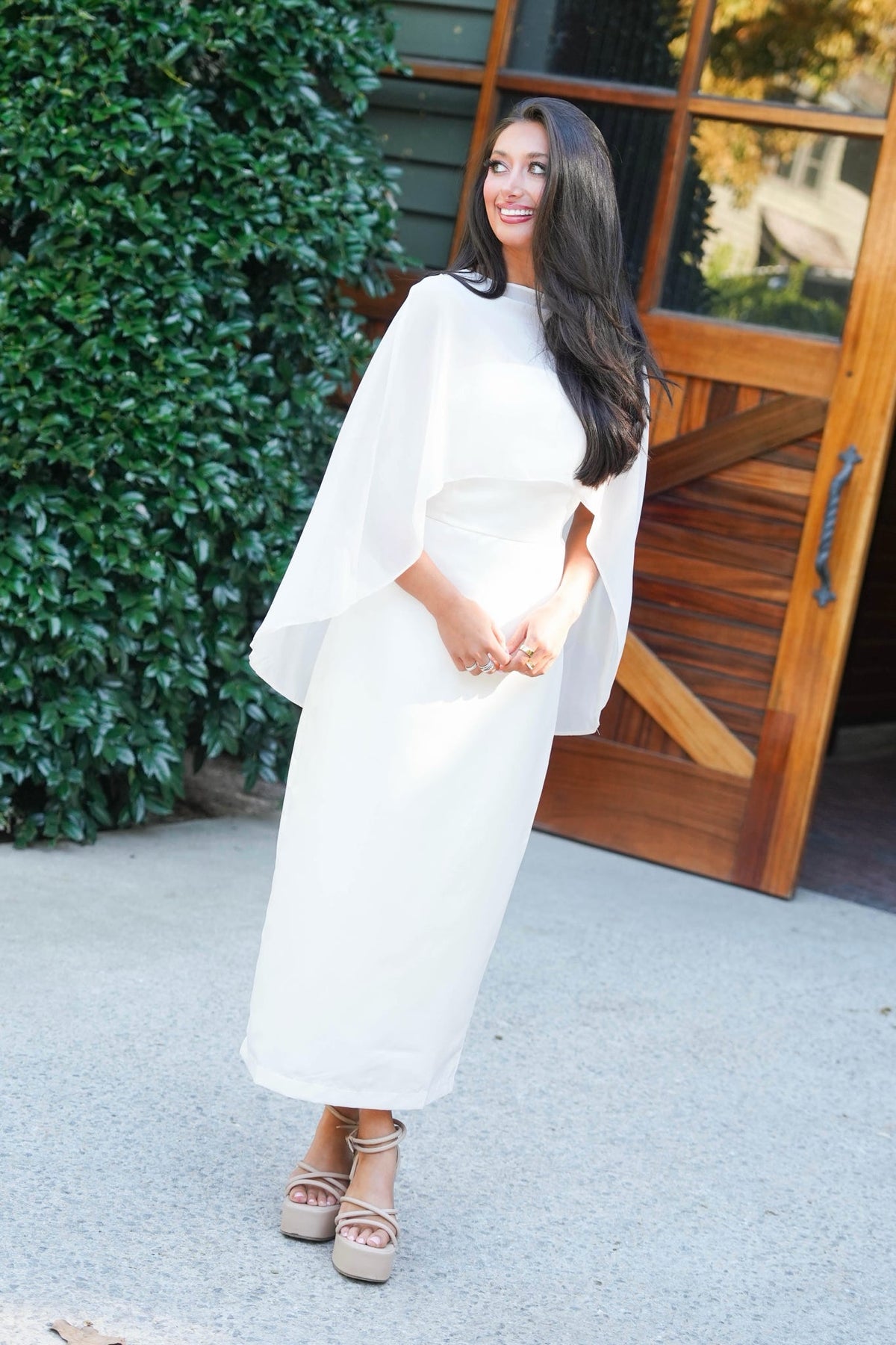 woman standing in front of barn door one wearing a white midi dress with sheer cape overlay