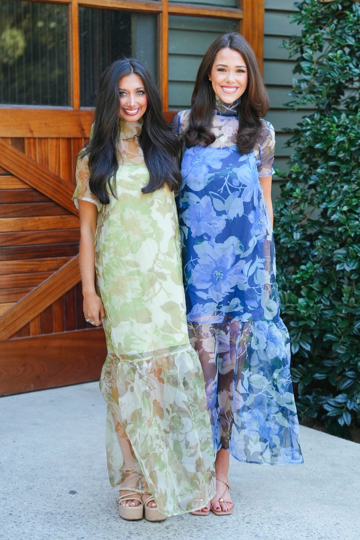 Two women standing outdoors in front of a wooden door and greenery, wearing long sheer maxi dresses with floral prints. One dress is light yellow-green, and the other is blue with a darker floral pattern.