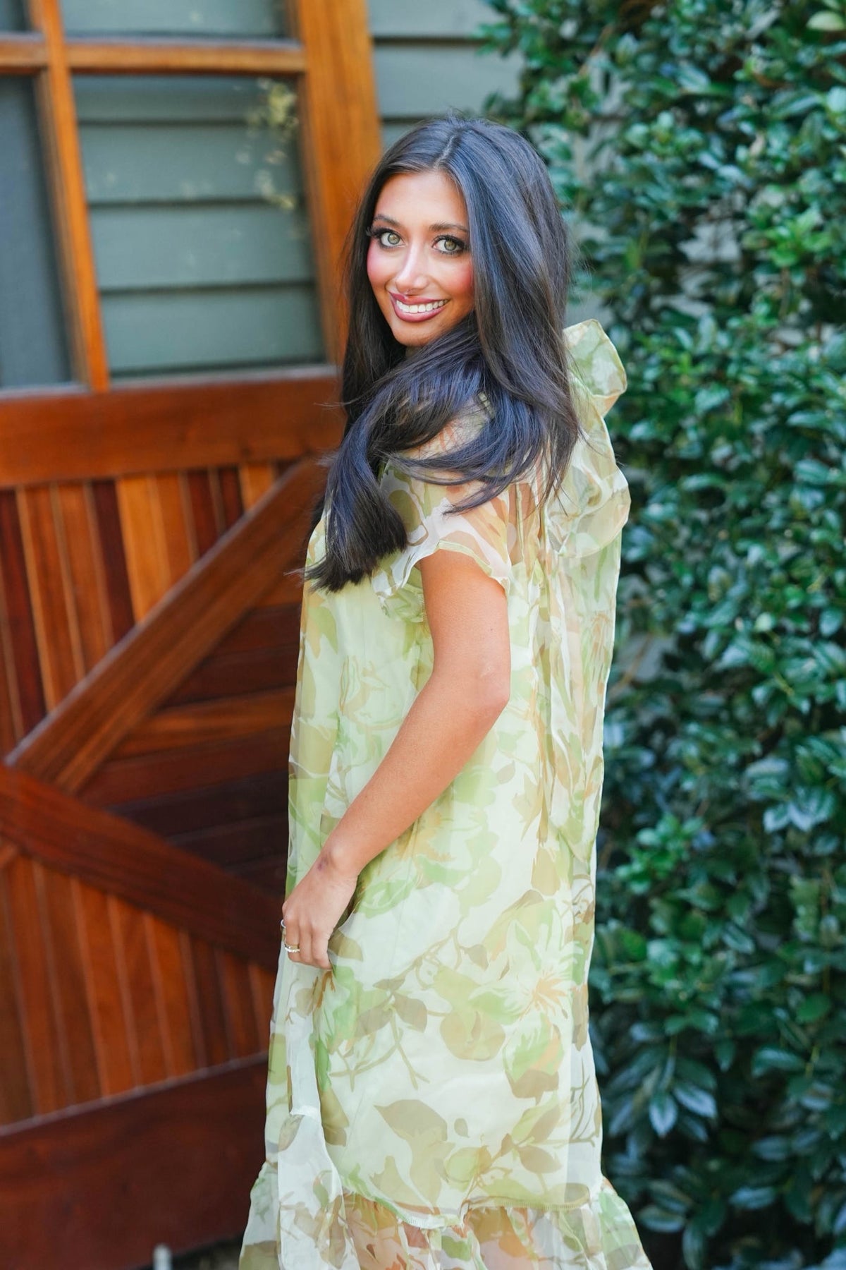A woman wearing a green floral midi dress with sheer sleeves and a sheer overlay skirt, standing outdoors in front of a wooden barn door and green foliage.