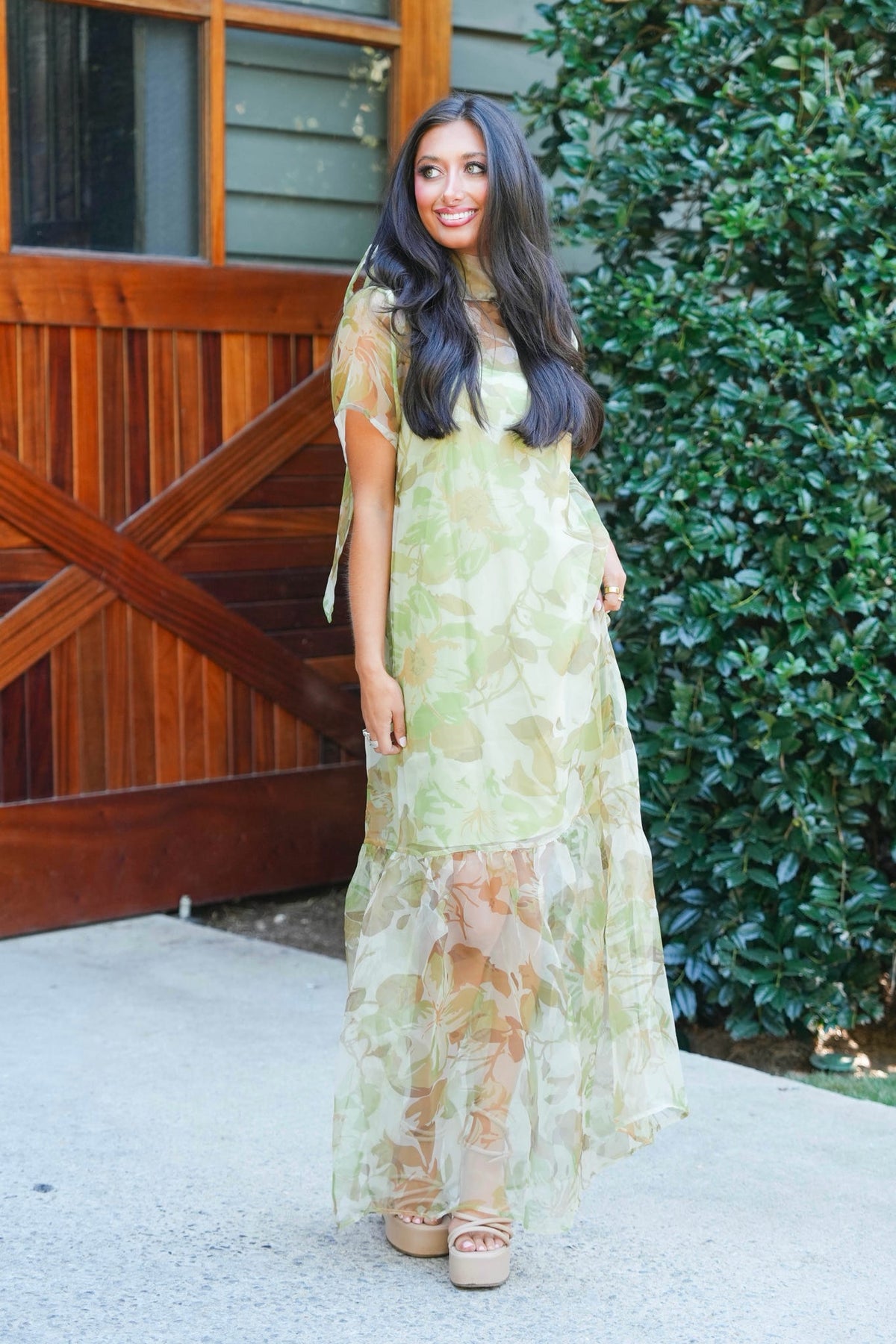 A woman wearing a sheer, light green and beige floral maxi dress with tie-shoulder details, standing outdoors in front of a wooden barn door and green foliage.