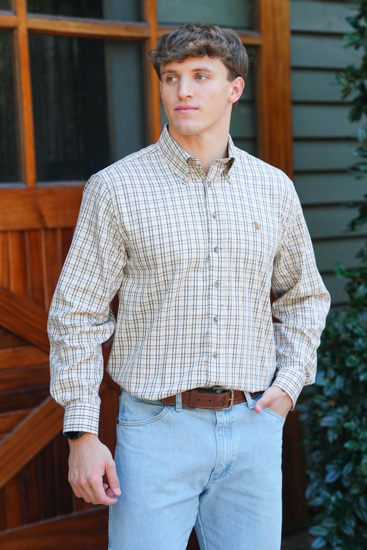 A man wearing a beige and brown plaid button-down shirt with light blue jeans, standing in front of a wooden door and house siding.