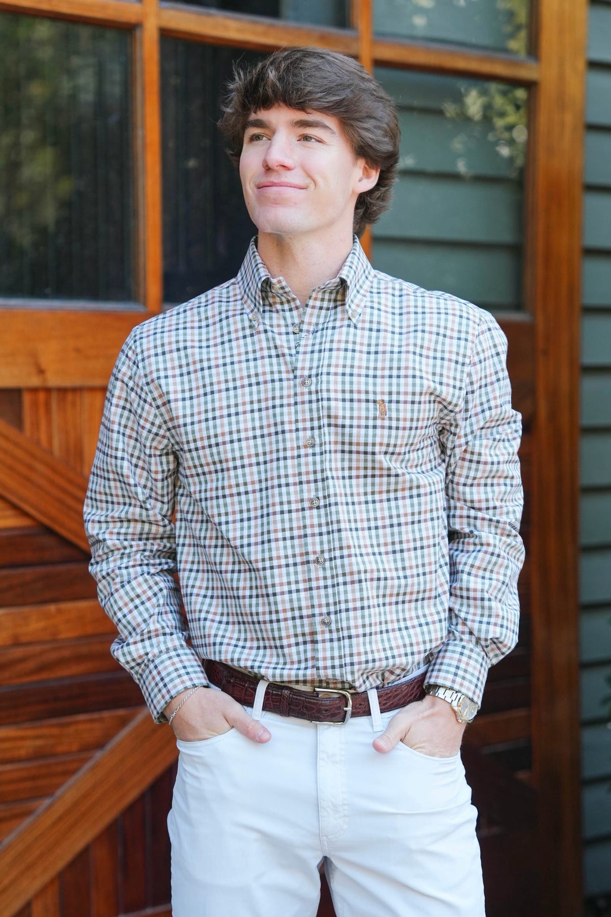 A young man wearing a long-sleeve button-down plaid shirt in beige, brown, and blue tones, paired with white pants and a brown belt, standing in front of a rustic wooden door.