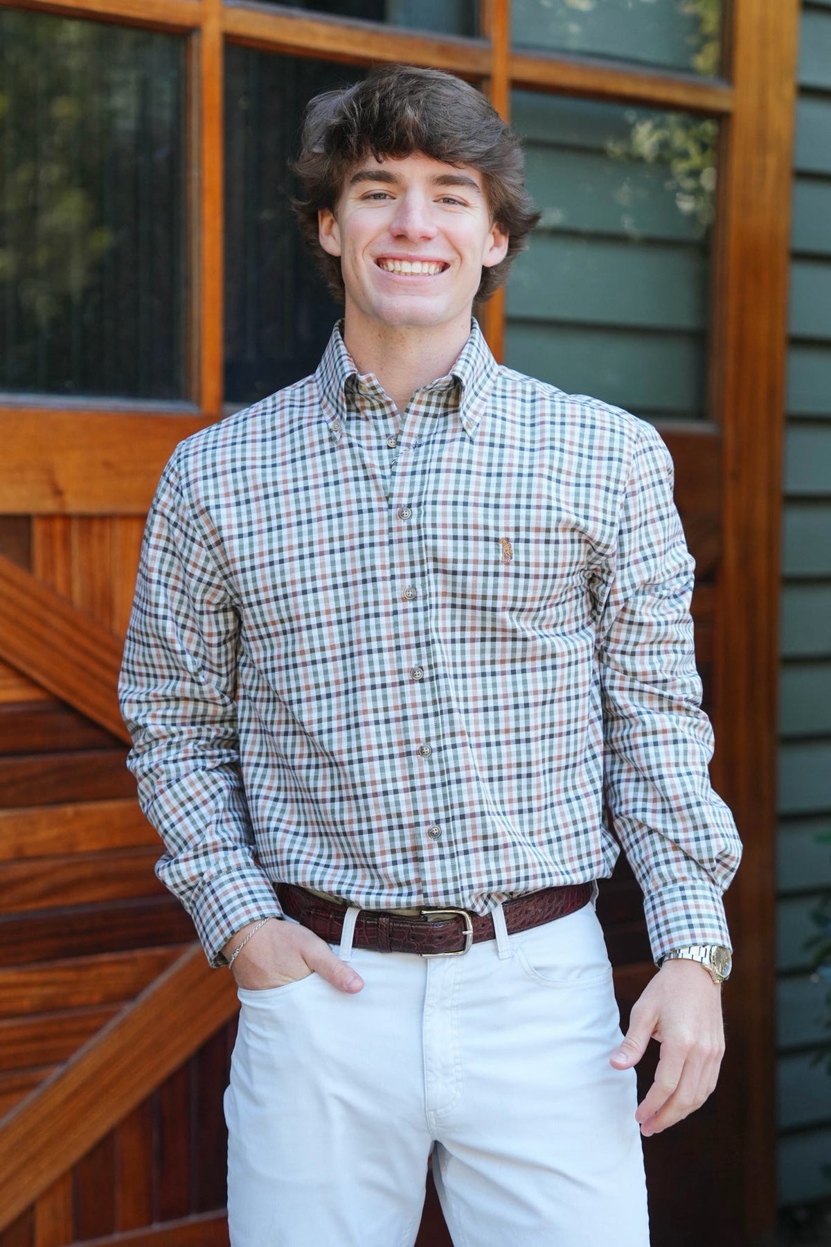 A smiling young man wearing a long-sleeve button-down plaid shirt in beige, blue, and brown, paired with white pants and a brown belt, standing in front of a rustic wooden barn door.