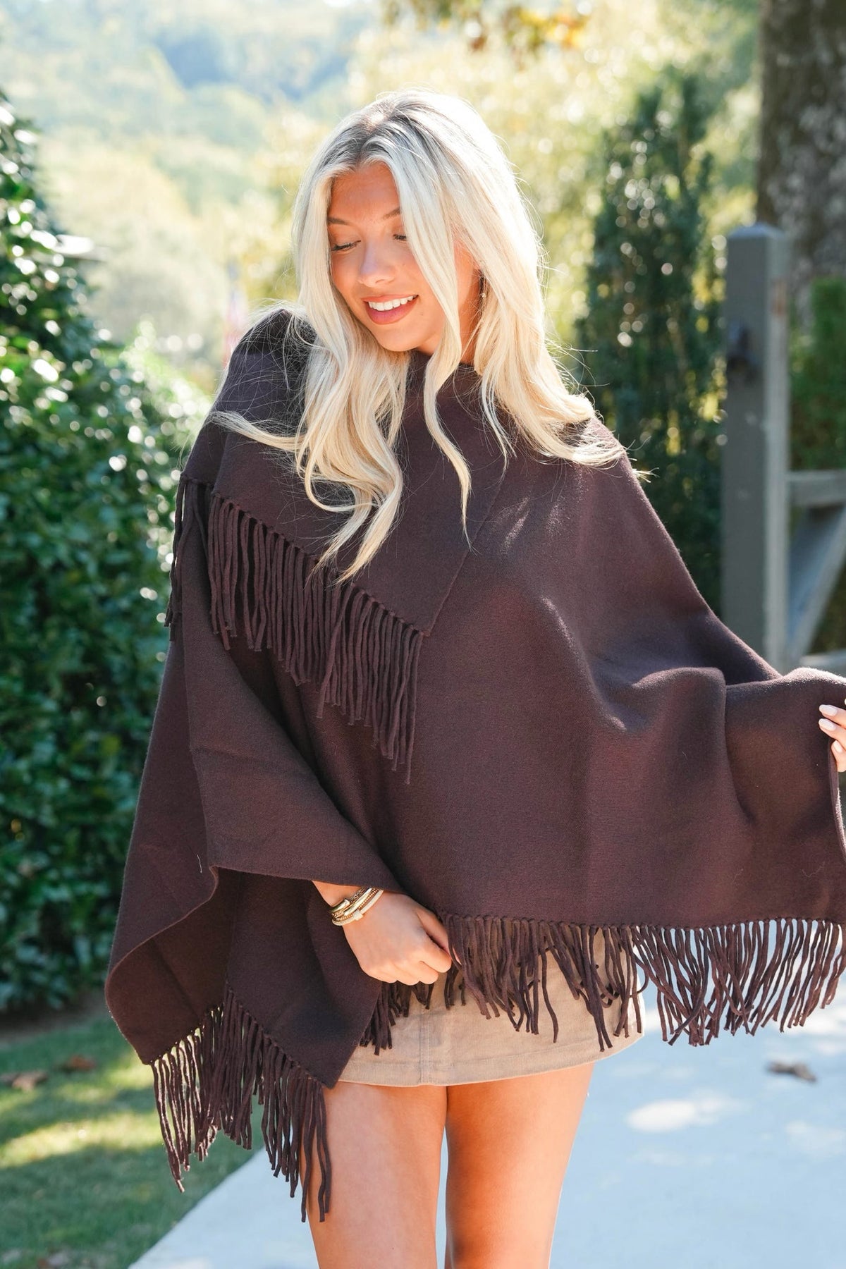 A woman wearing a dark brown fringed poncho in an outdoor garden setting.