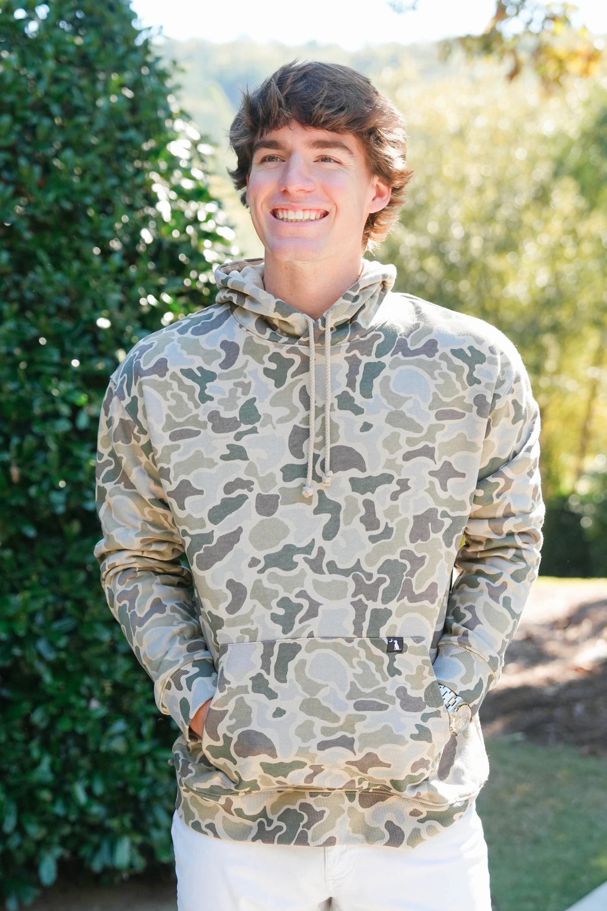 A smiling young man wearing a camouflage pullover hoodie with a kangaroo pocket, standing outdoors in front of a green hedge.