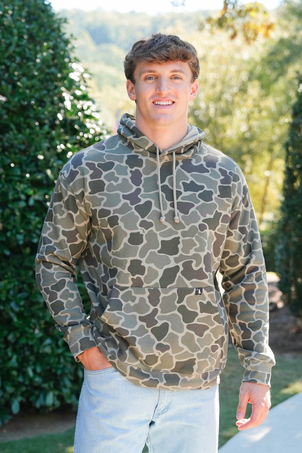 A man wearing a camouflage hoodie with a green and brown pattern, paired with light blue jeans, standing outdoors in front of a green hedge.