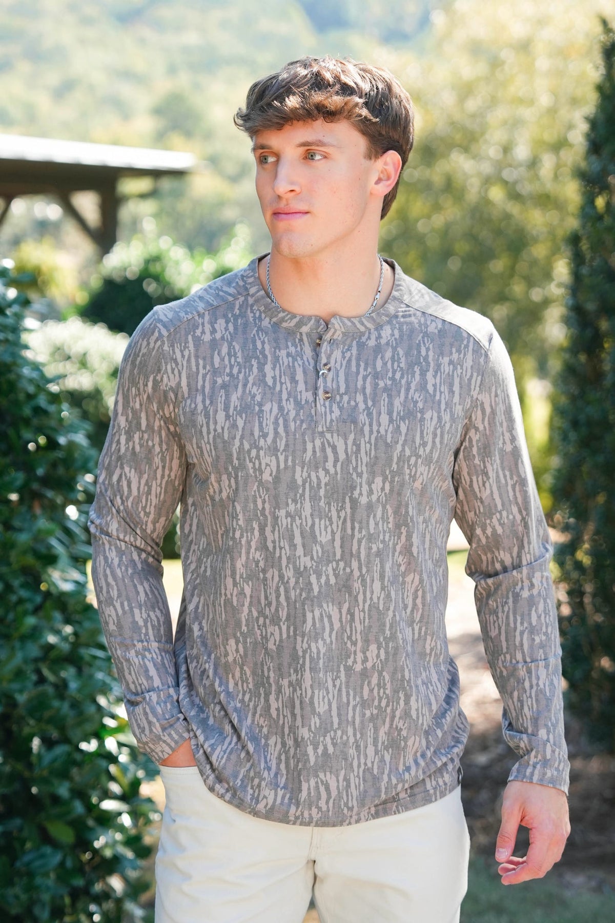 A man wearing a long-sleeve henley shirt with a gray and white abstract pattern, standing outdoors in a garden setting.