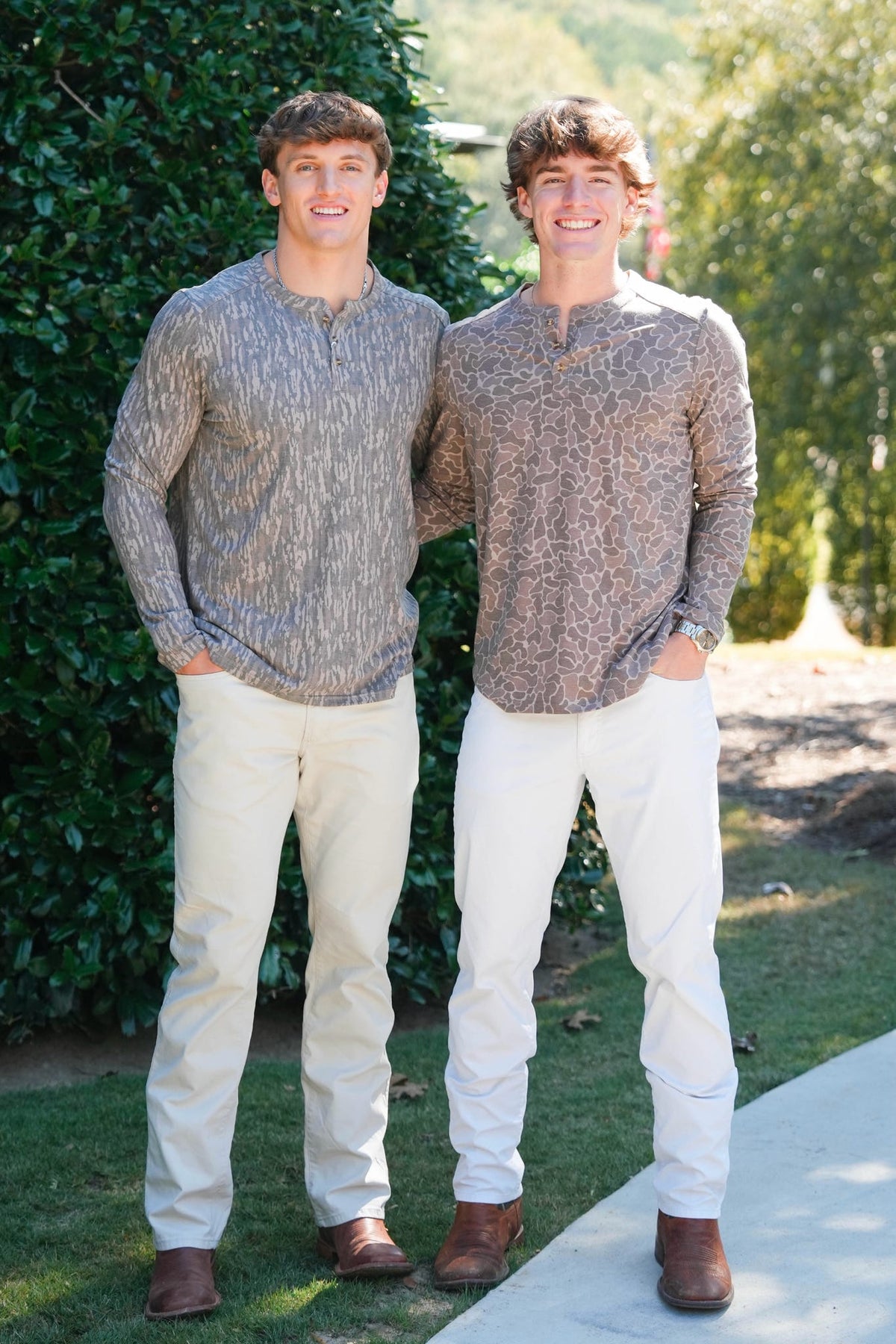 Two young men standing outdoors in front of green hedges, wearing matching long-sleeve henley shirts with a textured gray and brown abstract pattern, paired with off-white pants and brown shoes.