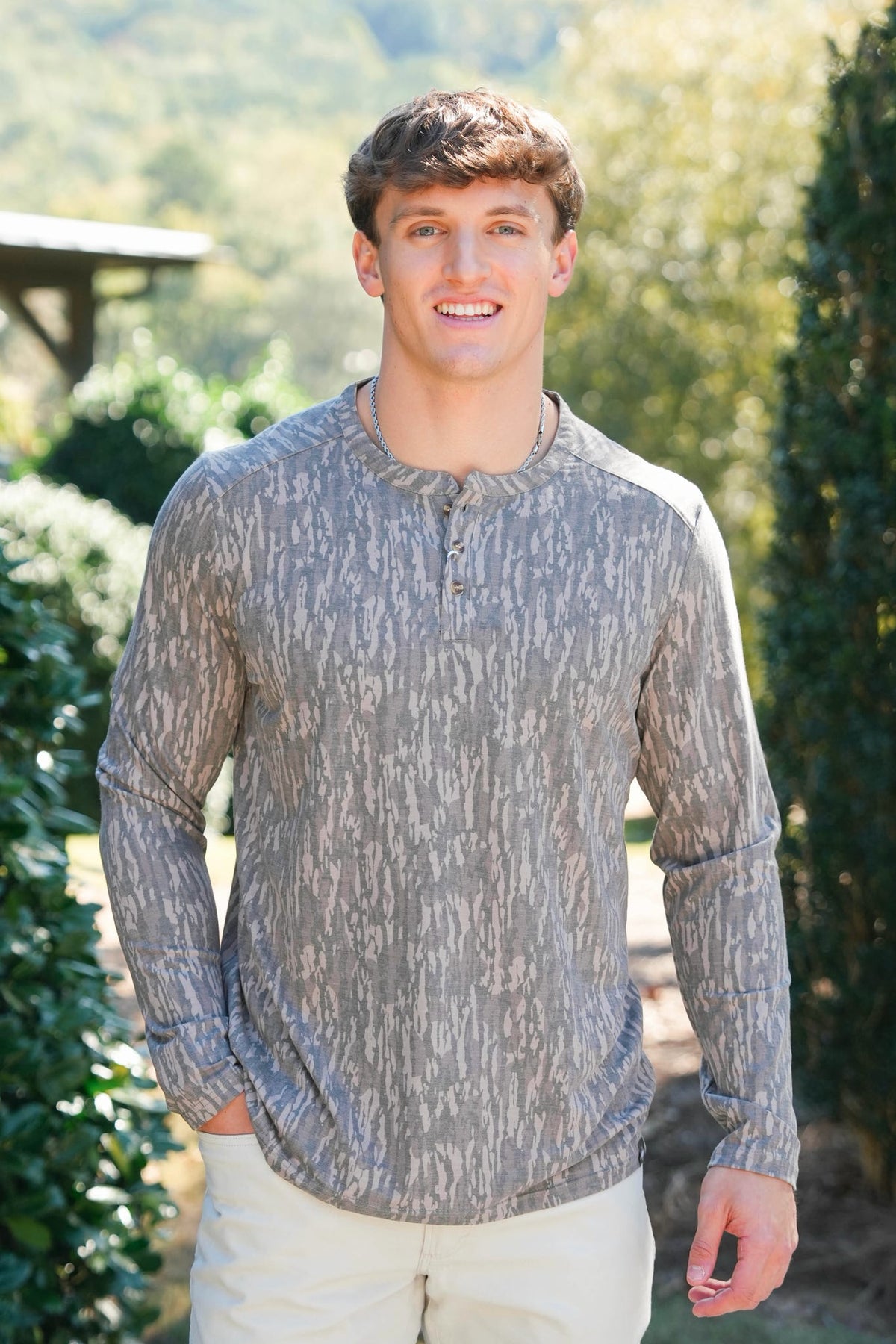 A smiling young man wearing a long-sleeve henley shirt with a gray and beige abstract pattern, standing outdoors in front of green trees and bushes.