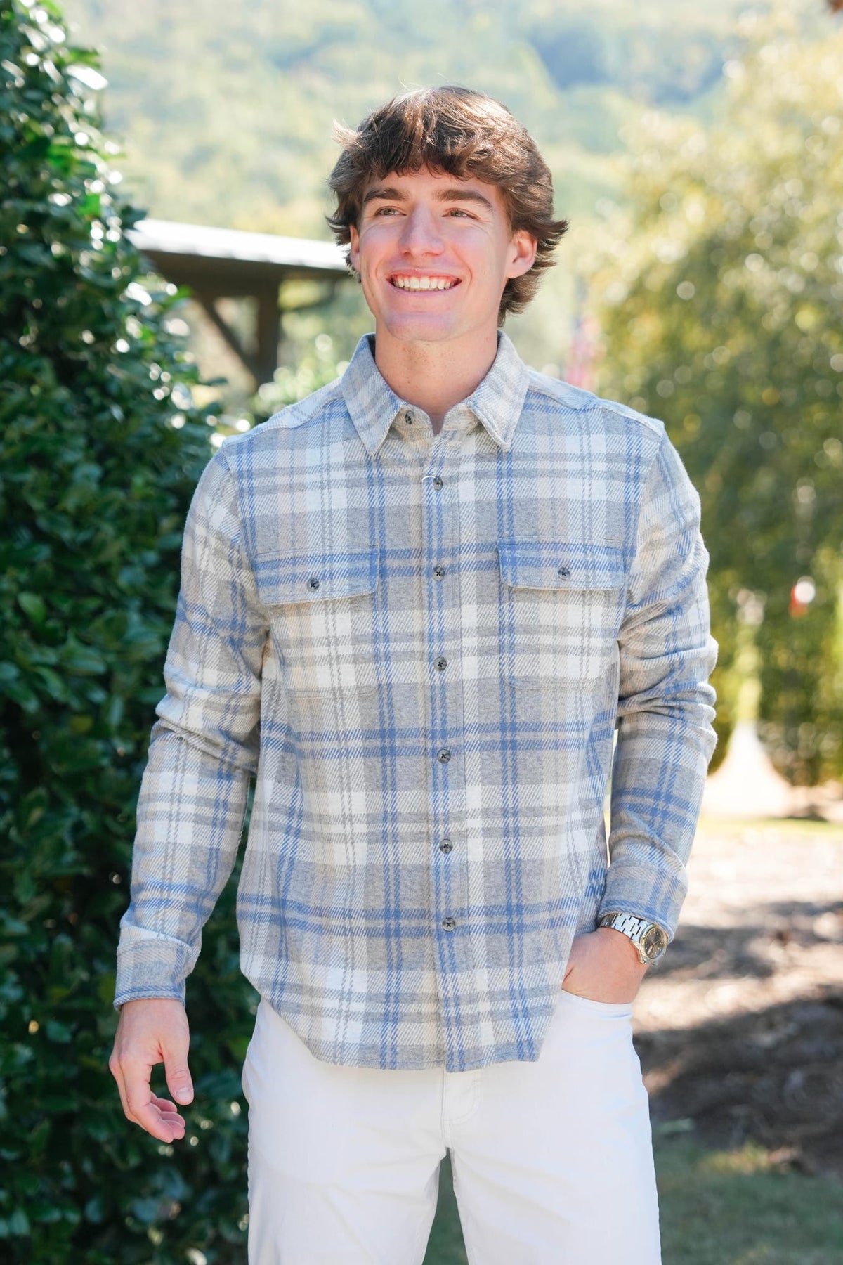 A smiling young man wearing a light blue and white plaid button-down shirt with white pants, standing outdoors in front of a green garden background.