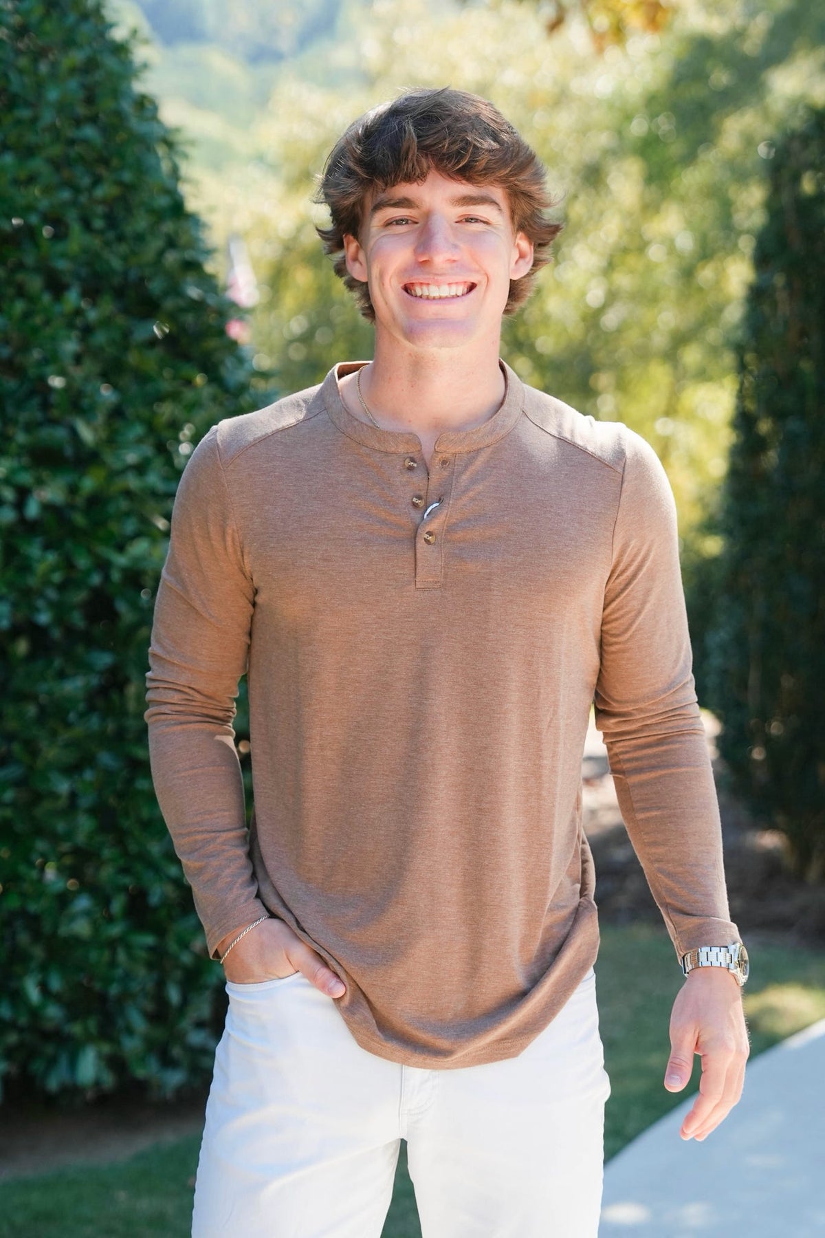 A smiling young man wearing a tan long-sleeve henley shirt and white pants, standing outdoors in front of green hedges and trees.