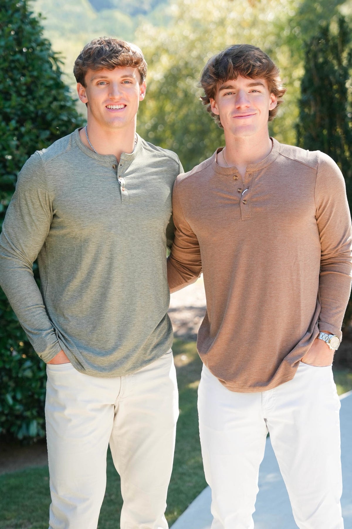Two men standing outdoors wearing long-sleeve henley shirts in muted sage green and taupe brown, paired with white pants.