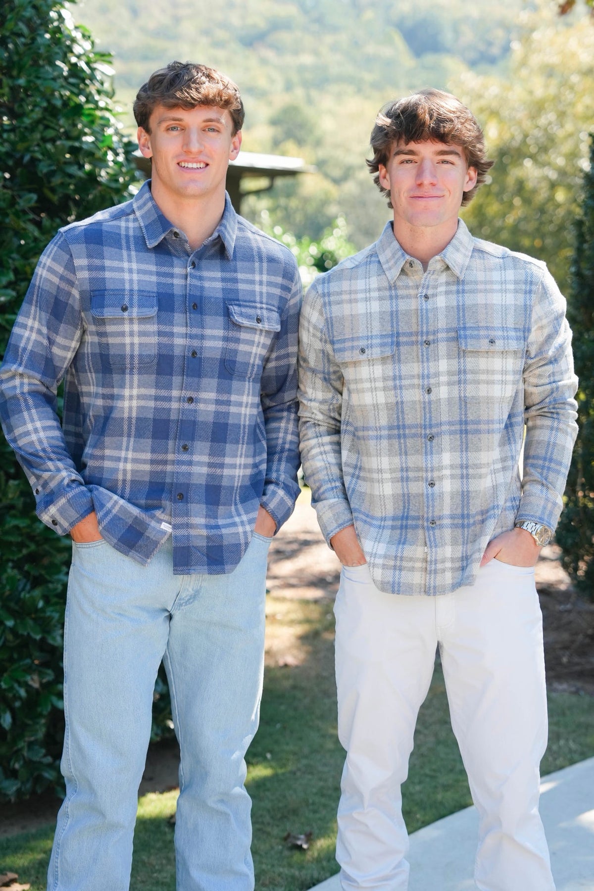 Two young men standing outdoors in front of a blurred green landscape, wearing long-sleeve plaid button-up shirts. The man on the left wears a medium blue and white plaid shirt with light blue jeans, and the man on the right wears a light blue and white plaid shirt with white jeans.