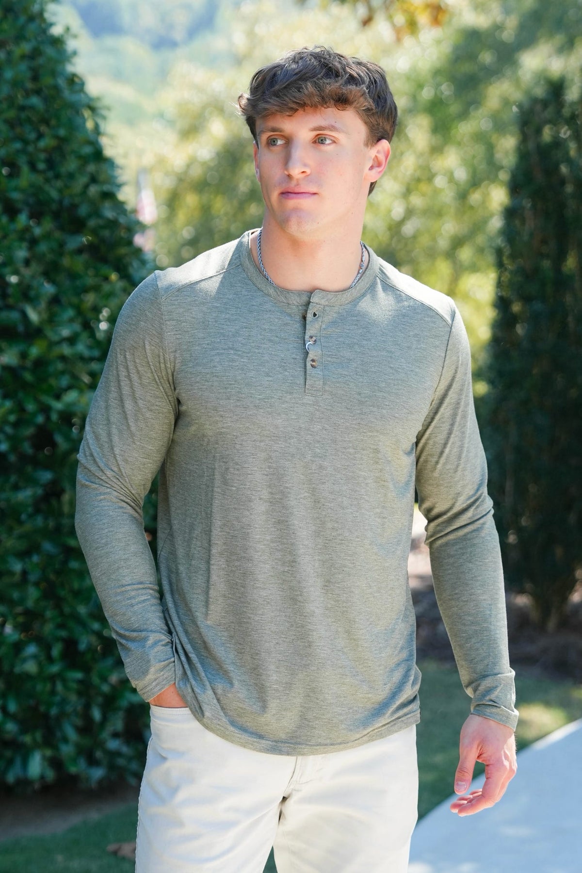 A man wearing a heather gray long-sleeve henley shirt and white pants, standing outdoors in front of green hedges and trees.