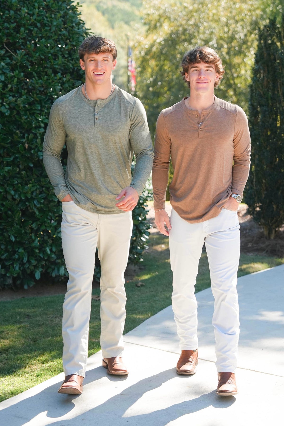 Two young men standing outdoors on a concrete path wearing long-sleeve henley shirts and white pants. The man on the left wears an olive green henley shirt, and the man on the right wears a tan/brown henley shirt.