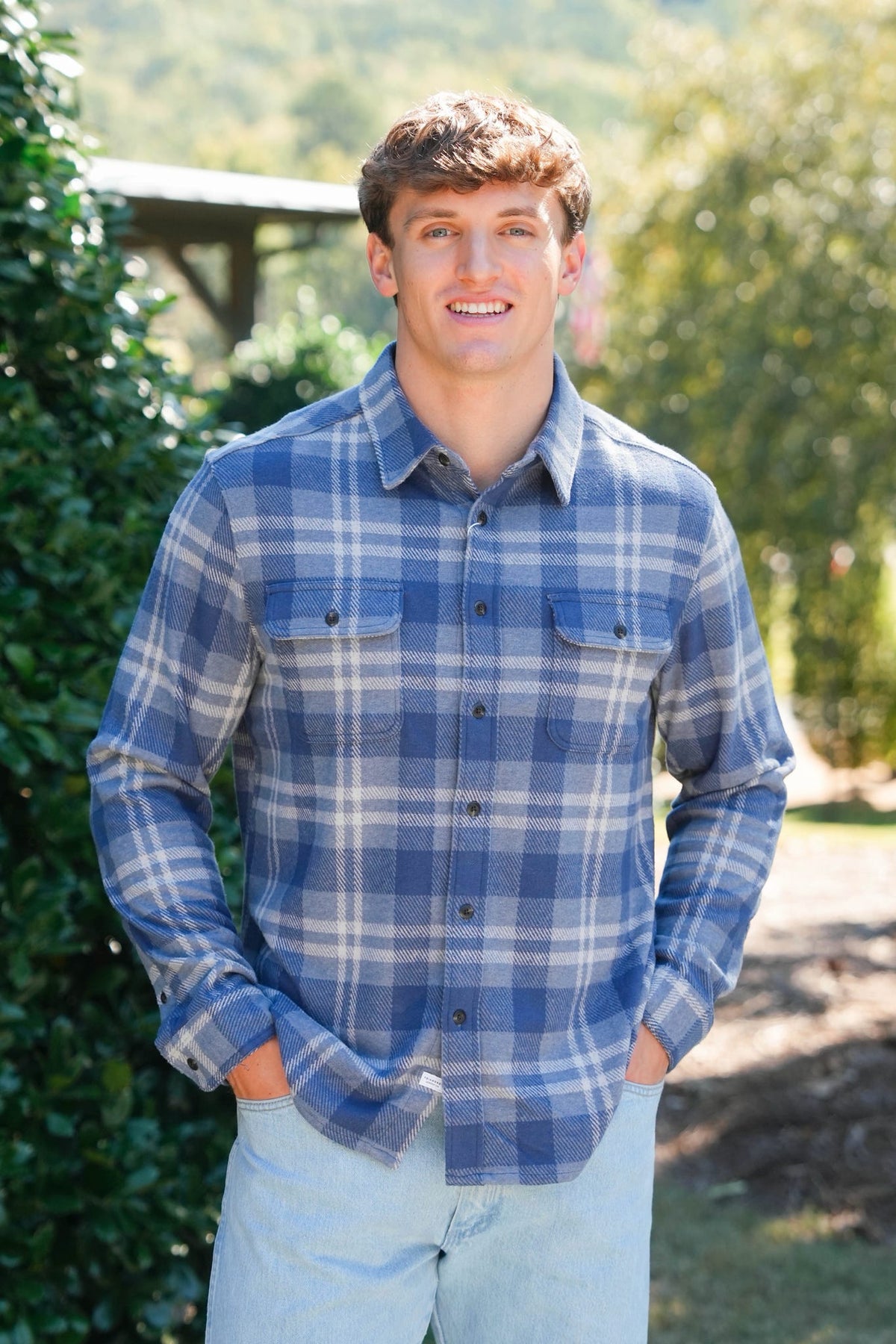 A young man smiling outdoors wearing a blue and white plaid long-sleeve button-down shirt and light blue jeans.
