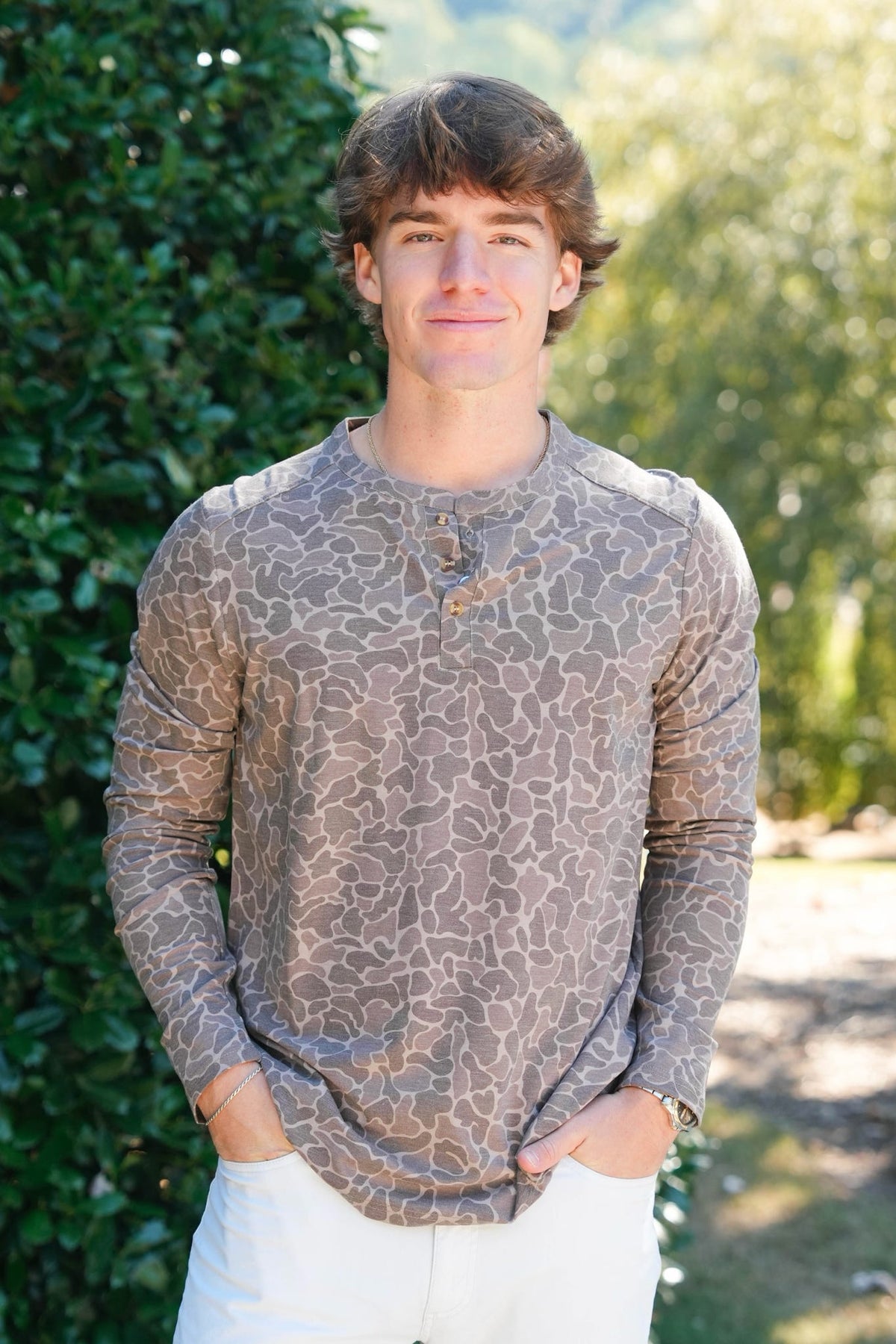 A young man wearing a brown and beige animal print long-sleeve henley shirt and white pants, standing outdoors in front of a green hedge.