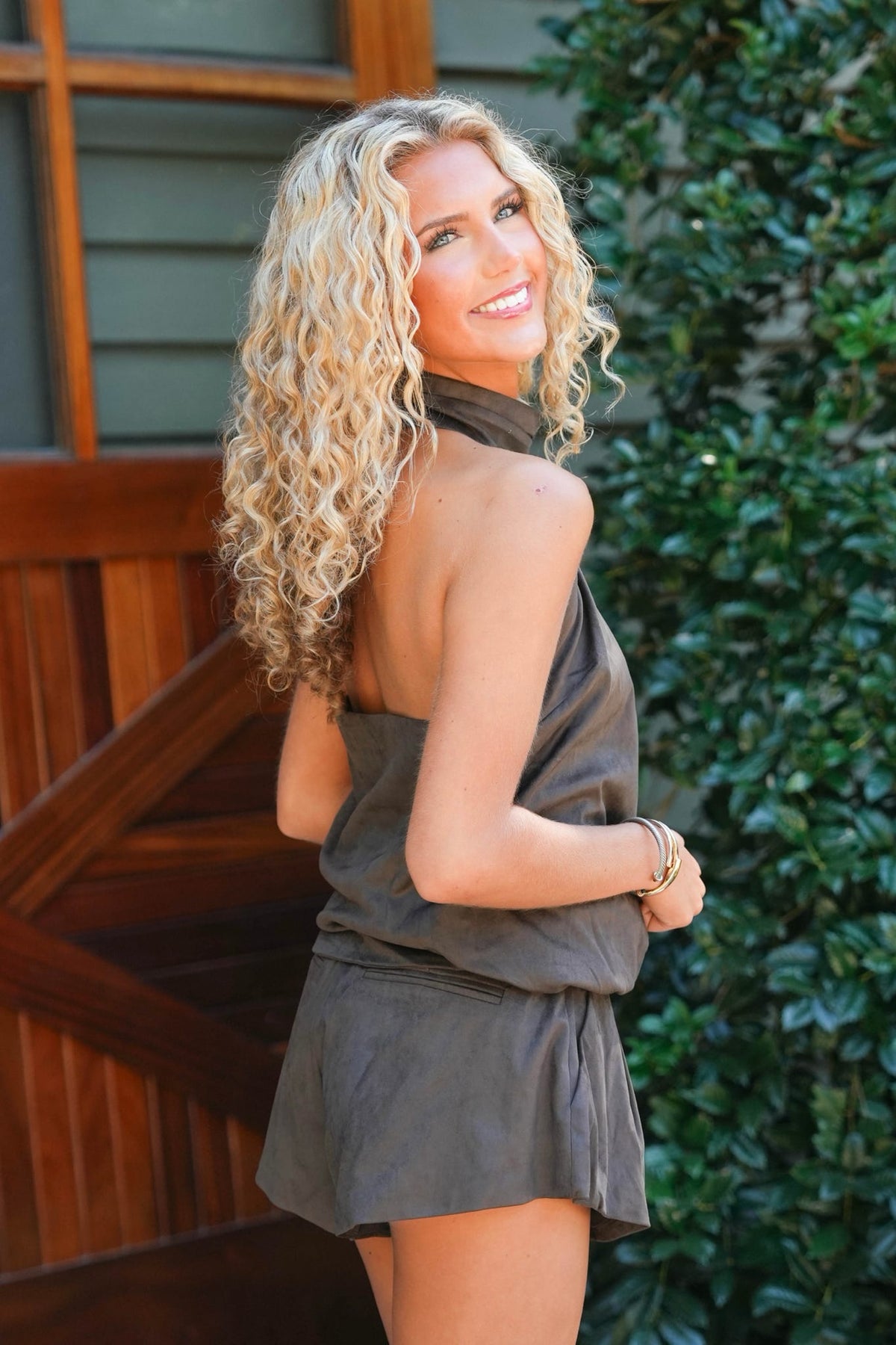 A woman with long curly blonde hair is shown from the back wearing a dark gray halter-neck sleeveless romper, standing outdoors in front of a wooden door and green foliage.